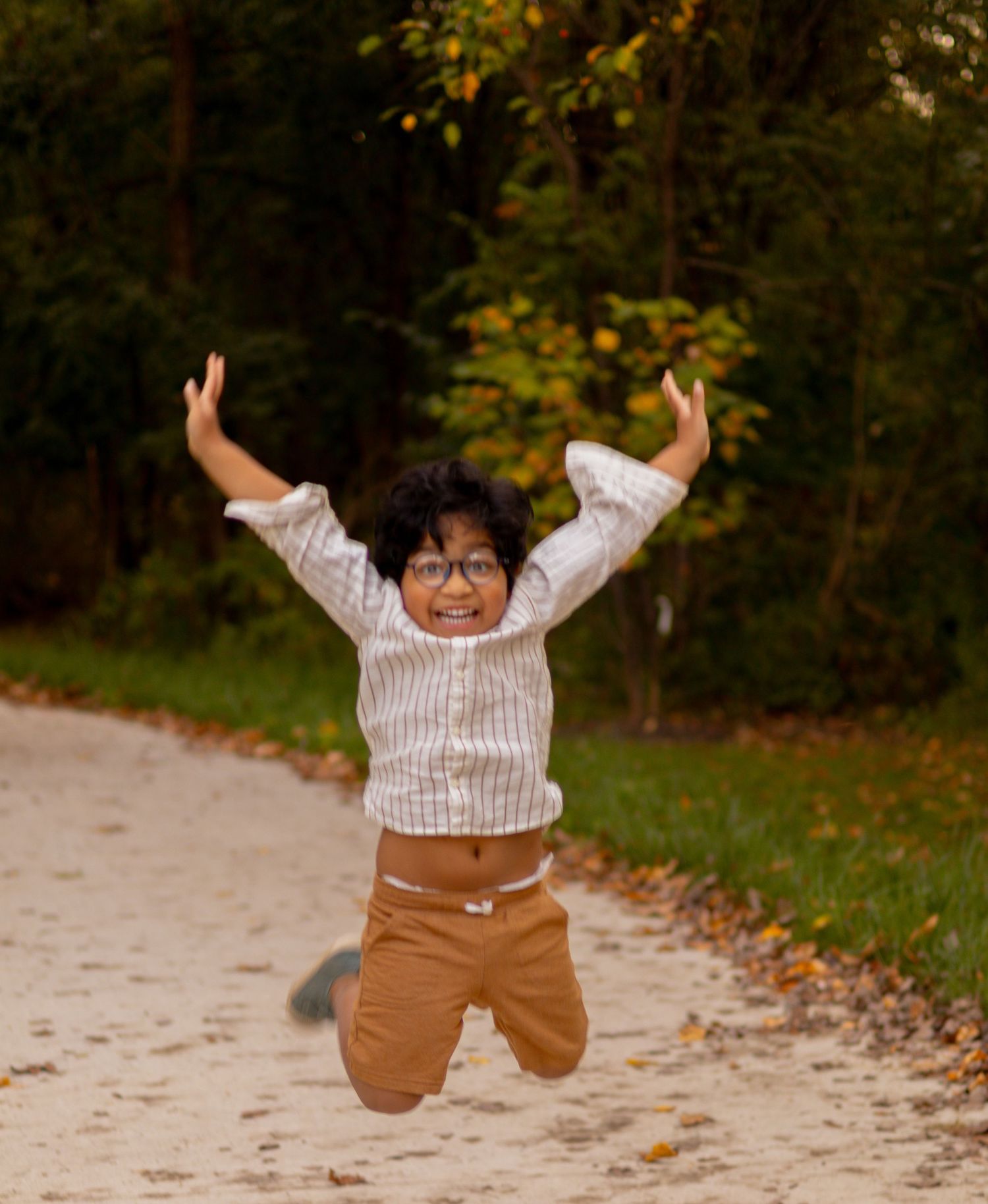 A child jumps with joy on a path wearing glasses and brown pants.