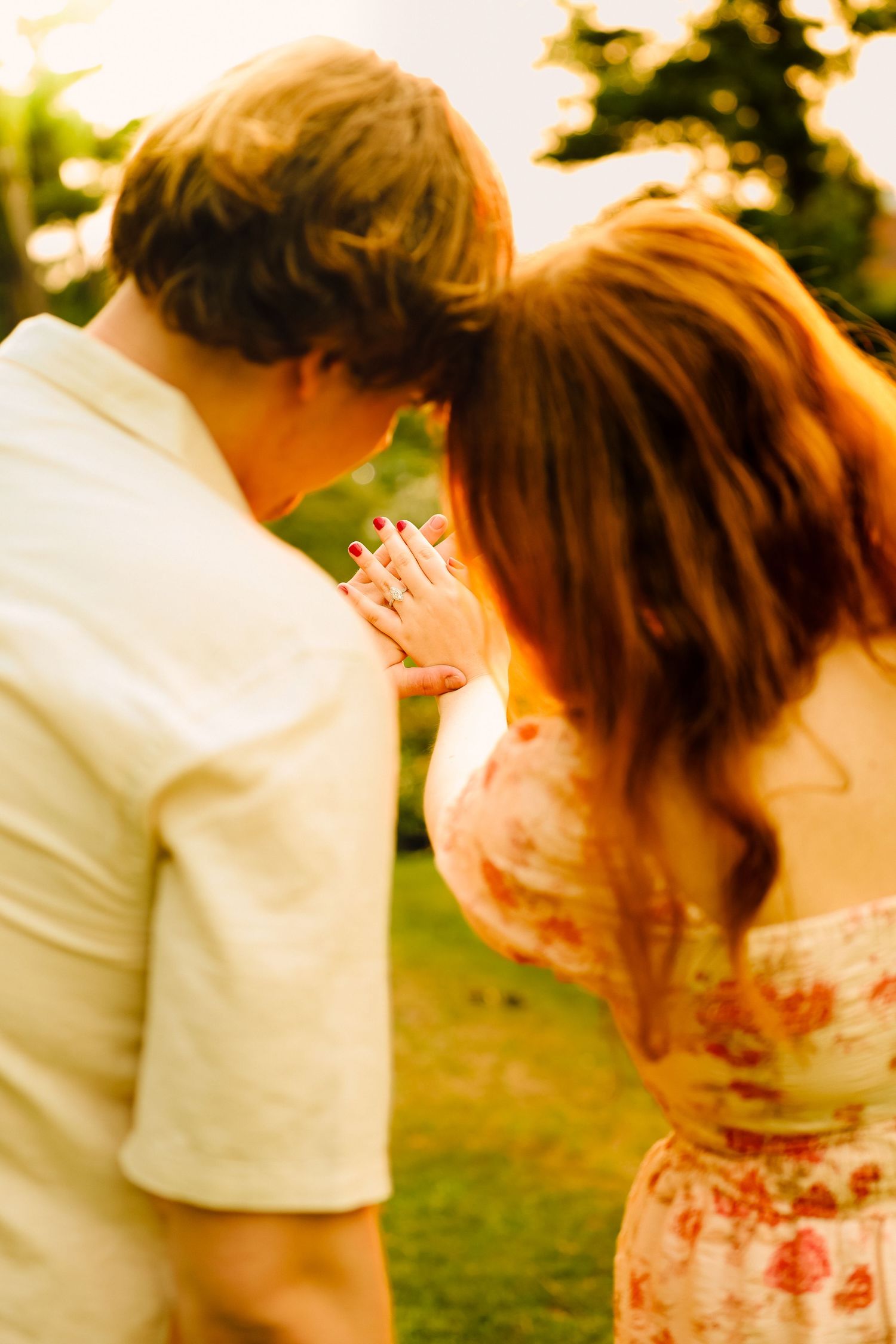 Couple sharing an intimate moment outdoors at golden hour, woman in floral dress showing engagement ring.