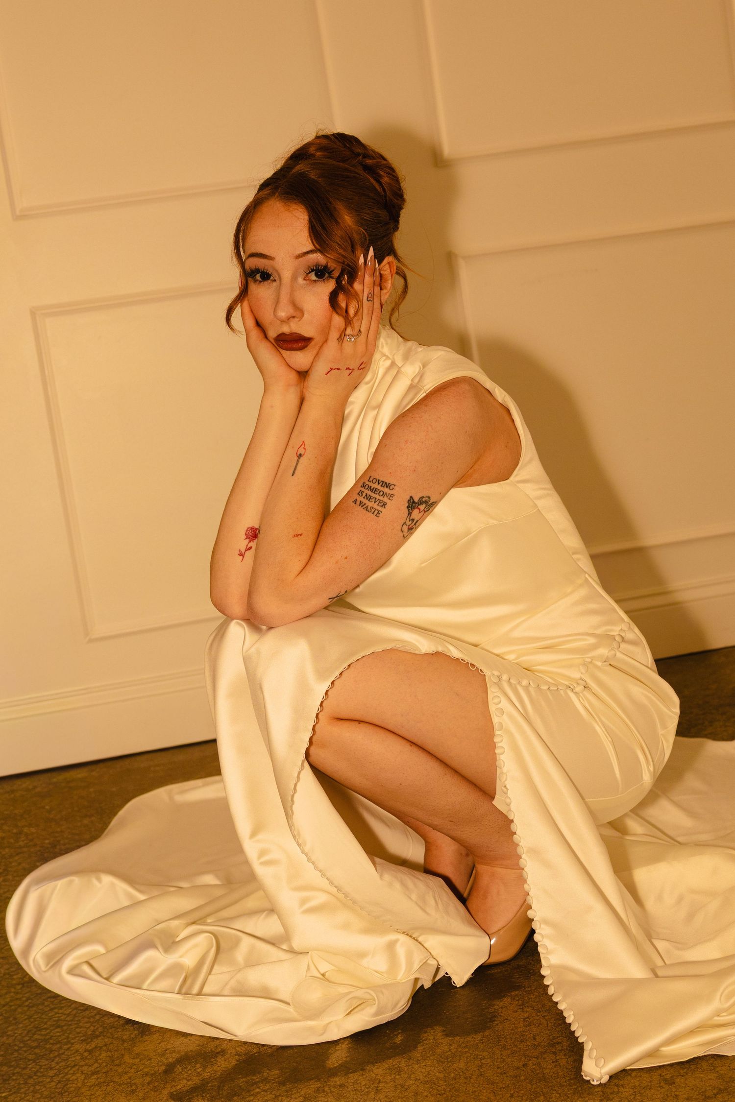 Woman in white satin dress sitting on floor, resting chin on hands, with tattoos visible, dark lips, updo hairstyle.