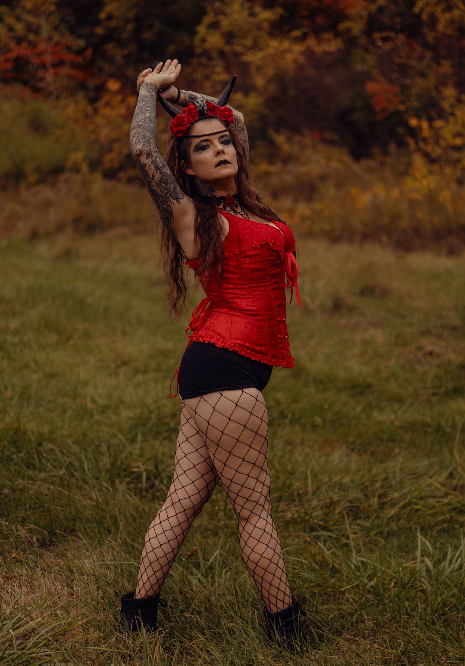 Woman in red corset, fishnet tights, devil horns headpiece, and tattoos poses outdoors in autumn setting.