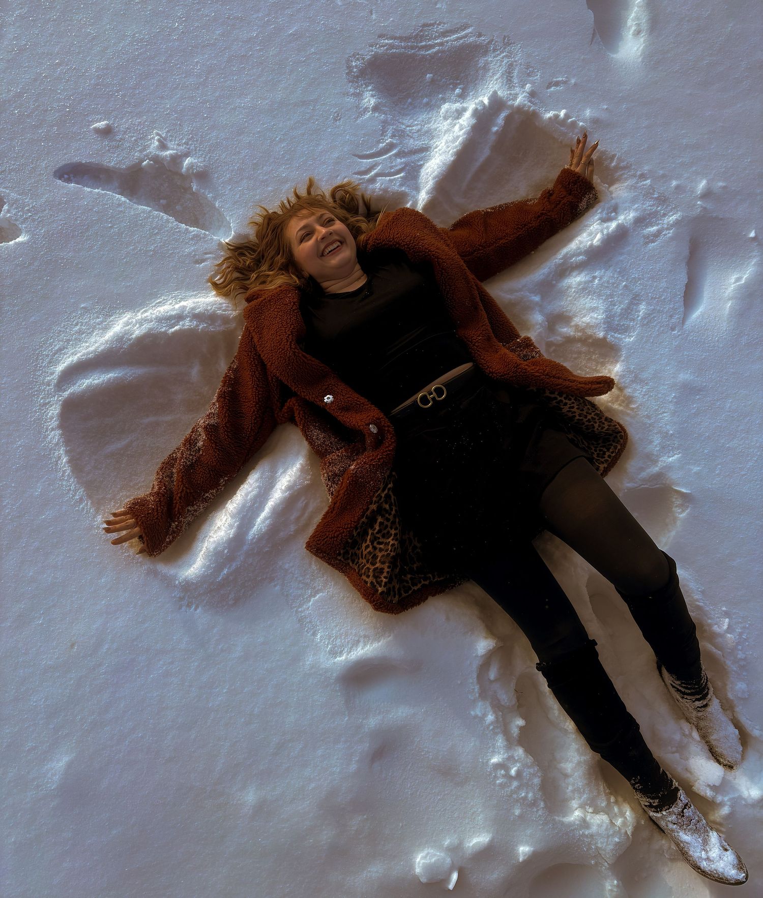 Woman in brown jacket making a snow angel, lying in white snow smiling, aerial view winter outdoor photo.