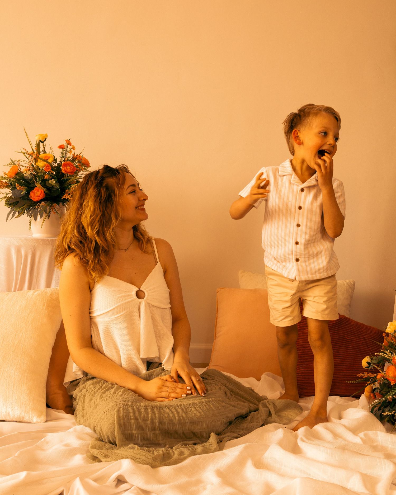 A woman sits cross-legged on a bed while a young boy stands beside her eating, with flowers in the background.