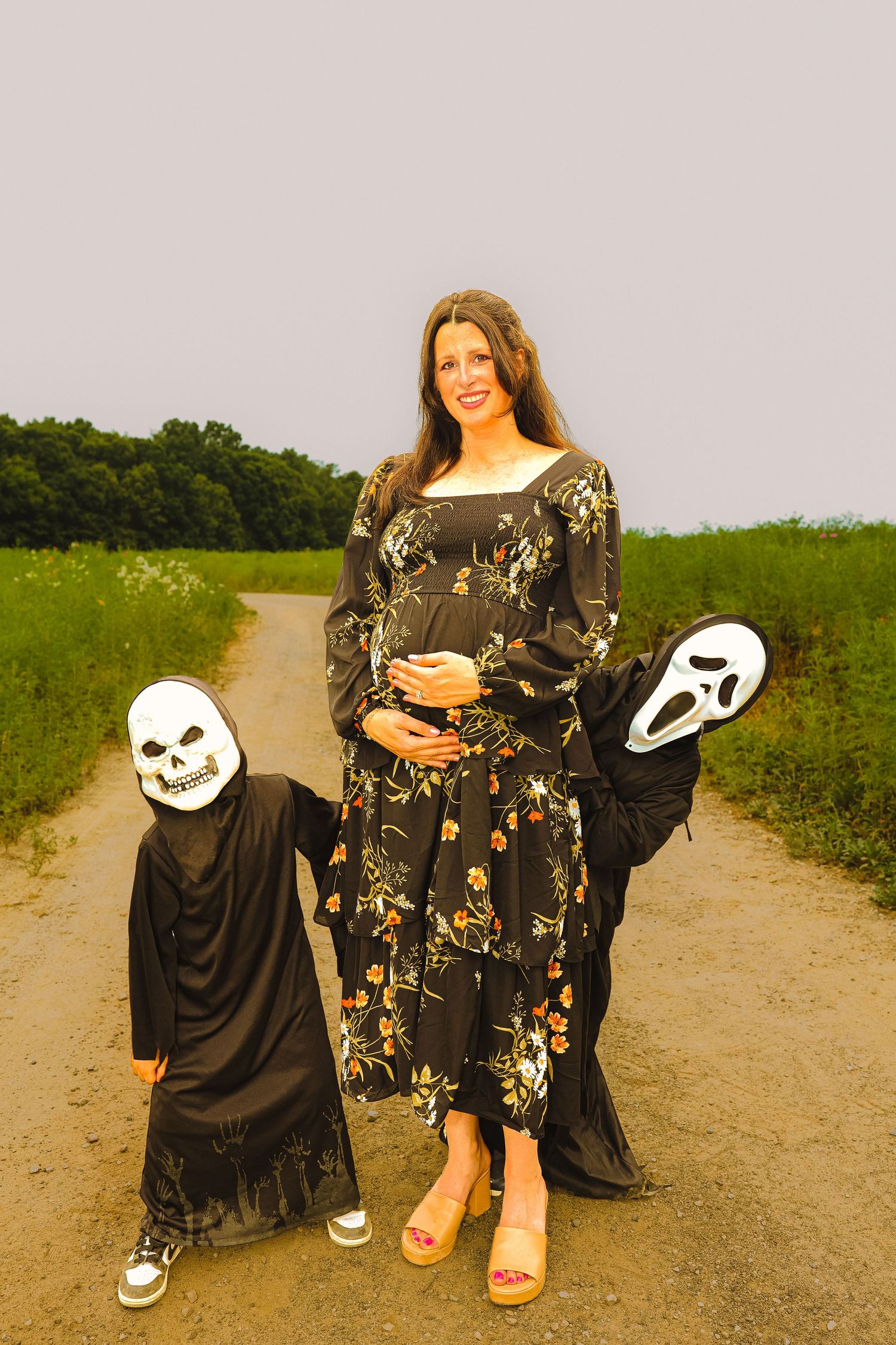 Pregnant woman in floral dress poses with two children in Halloween skeleton and ghost costumes on a rural dirt road.