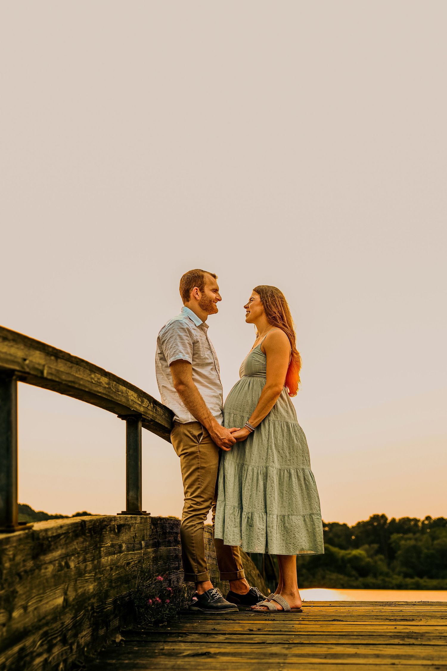 Couple holding hands on wooden bridge at sunset, woman in green dress with baby bump, man in casual shirt smiling at each other.