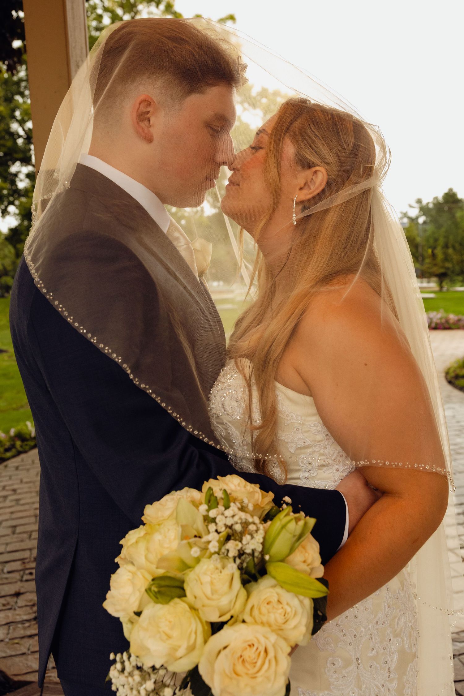 Bride and groom share an intimate moment under a sheer beaded veil, holding a bouquet of white roses in a sunlit garden.