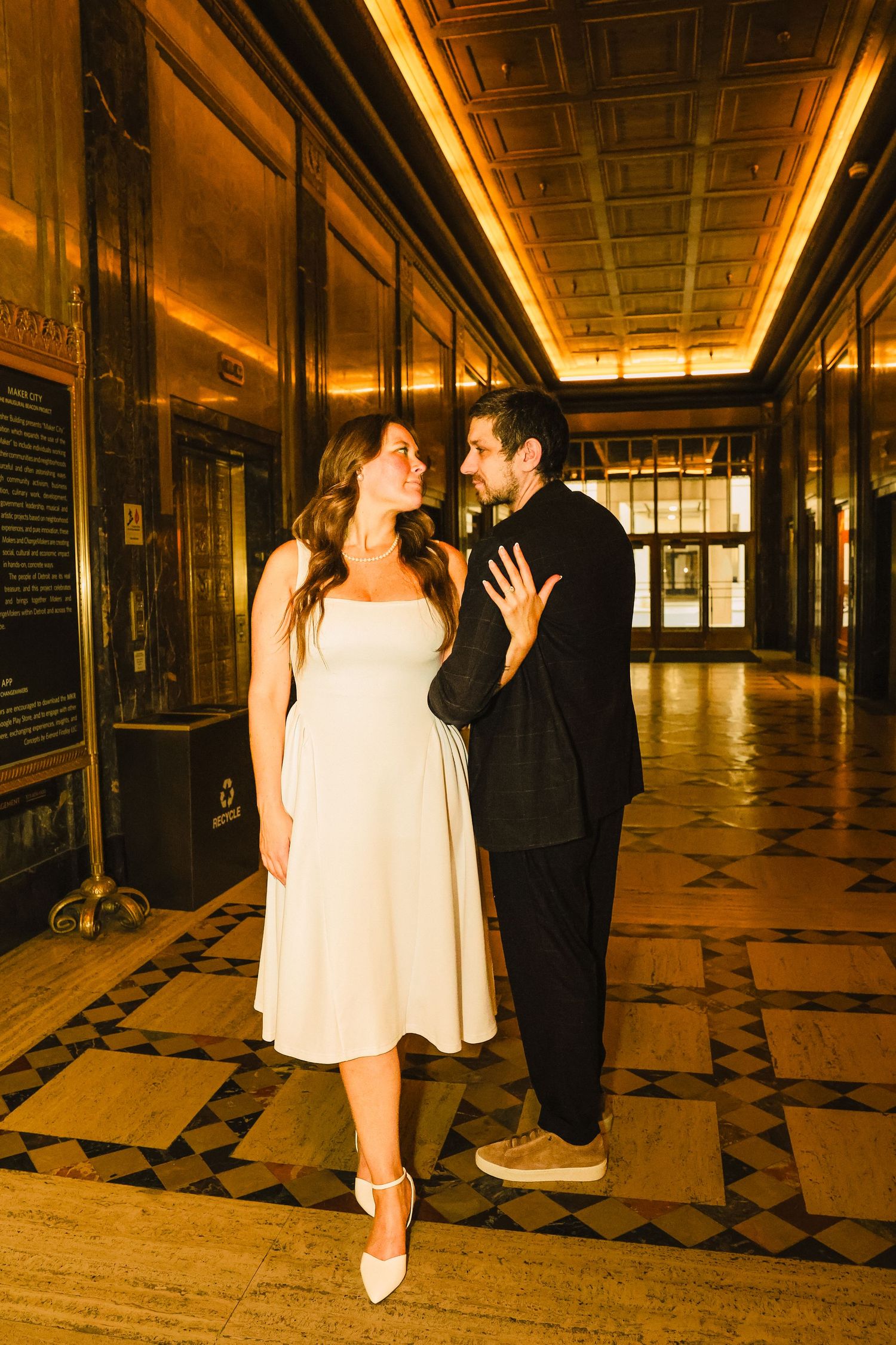 Couple in elegant hallway, woman in white midi dress and man in black suit, sharing a romantic moment under warm golden lights.