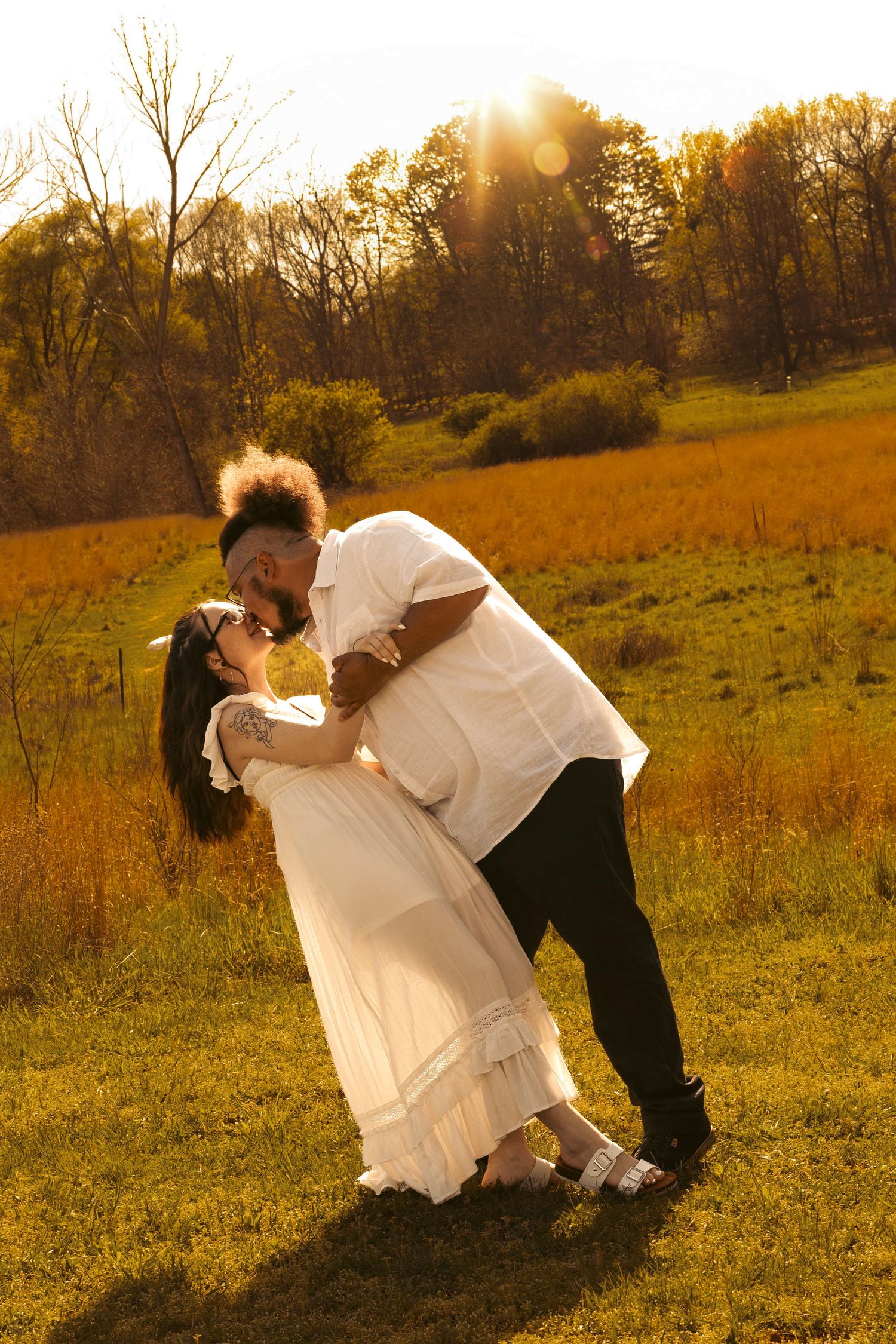 Couple sharing a romantic dip kiss in a golden sunlit field during sunset, woman in white dress, man in white shirt.