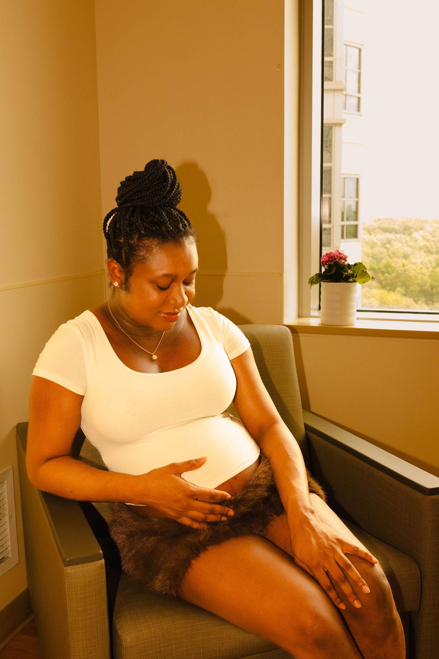 Pregnant Black woman sitting in chair touching her baby bump, wearing white top and brown skirt near window.