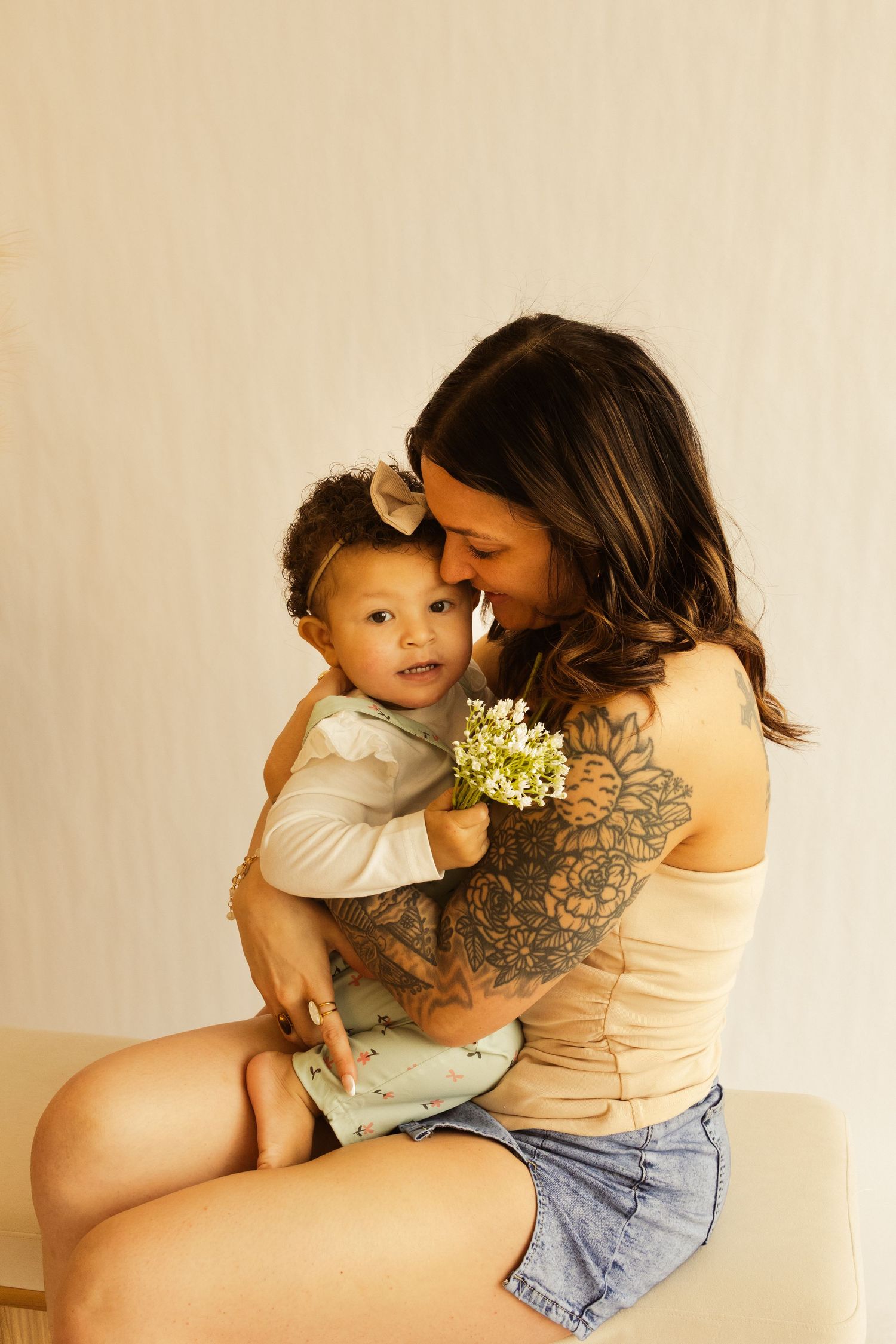 A tattooed mother tenderly nuzzles her smiling baby girl holding small white flowers while seated together.