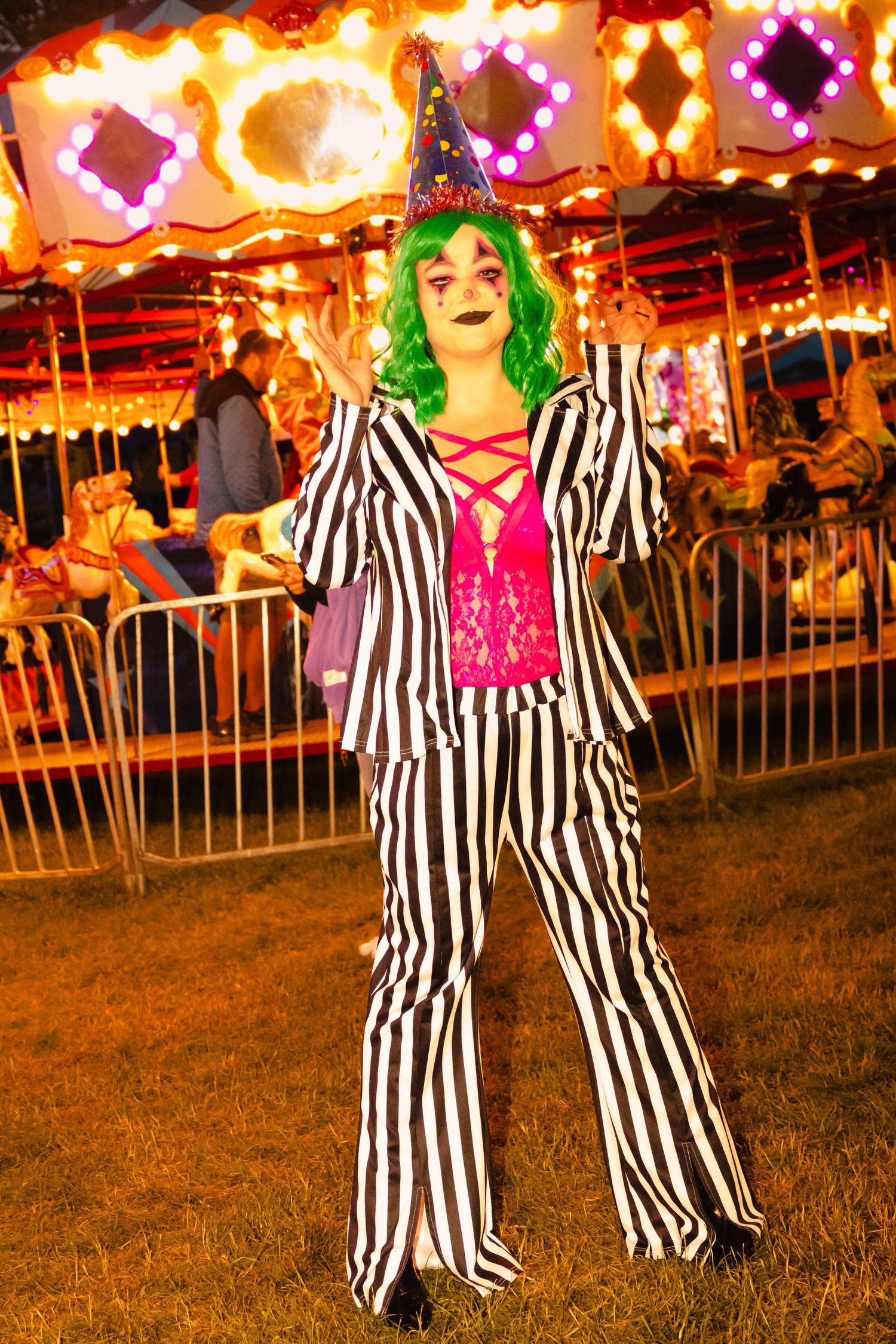 Person in striped suit, green wig, and witch hat poses at a carnival with a lit carousel in the background.