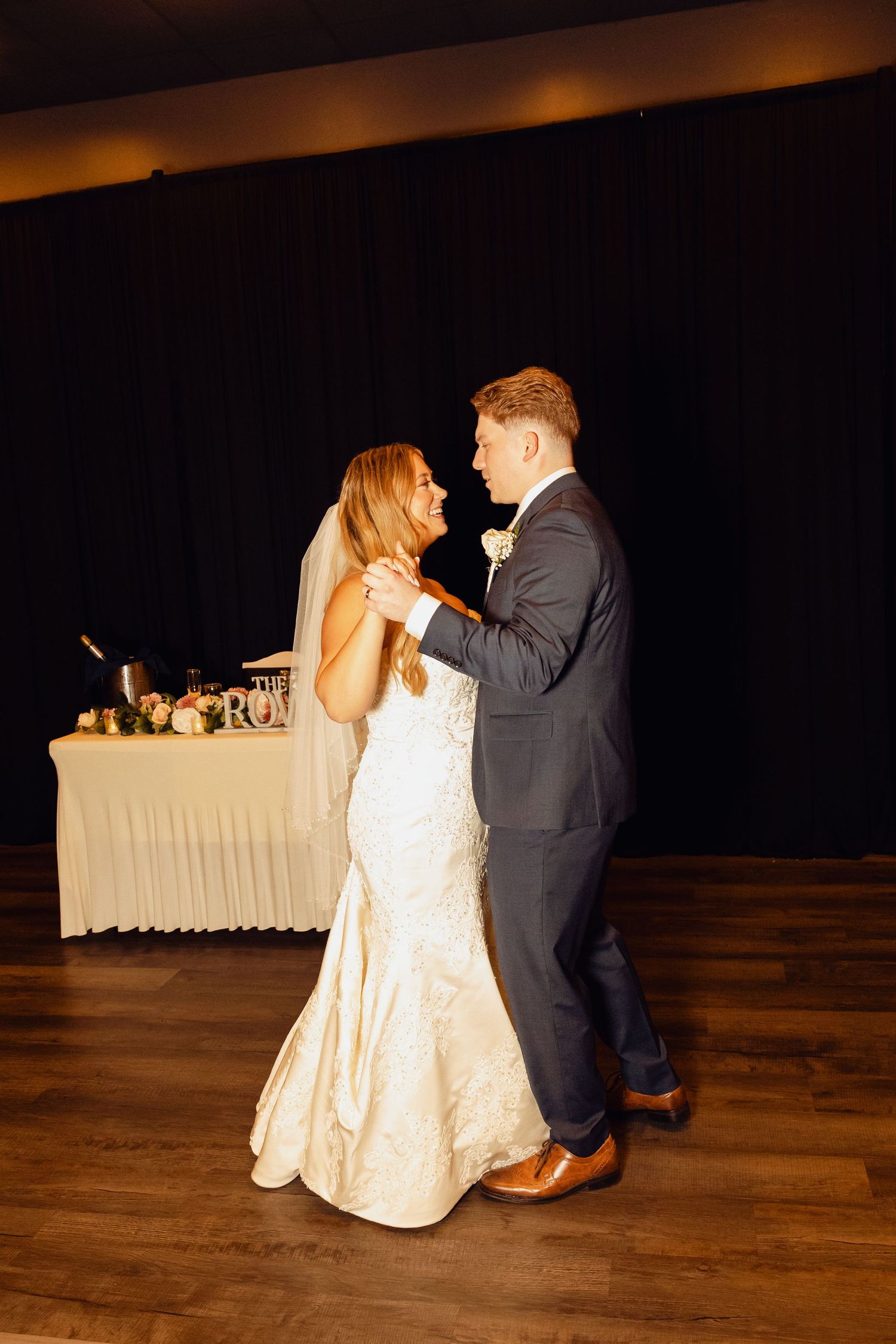 Bride in a lace mermaid gown and veil shares a first dance with the groom in a gray suit on a wood floor at a wedding reception.