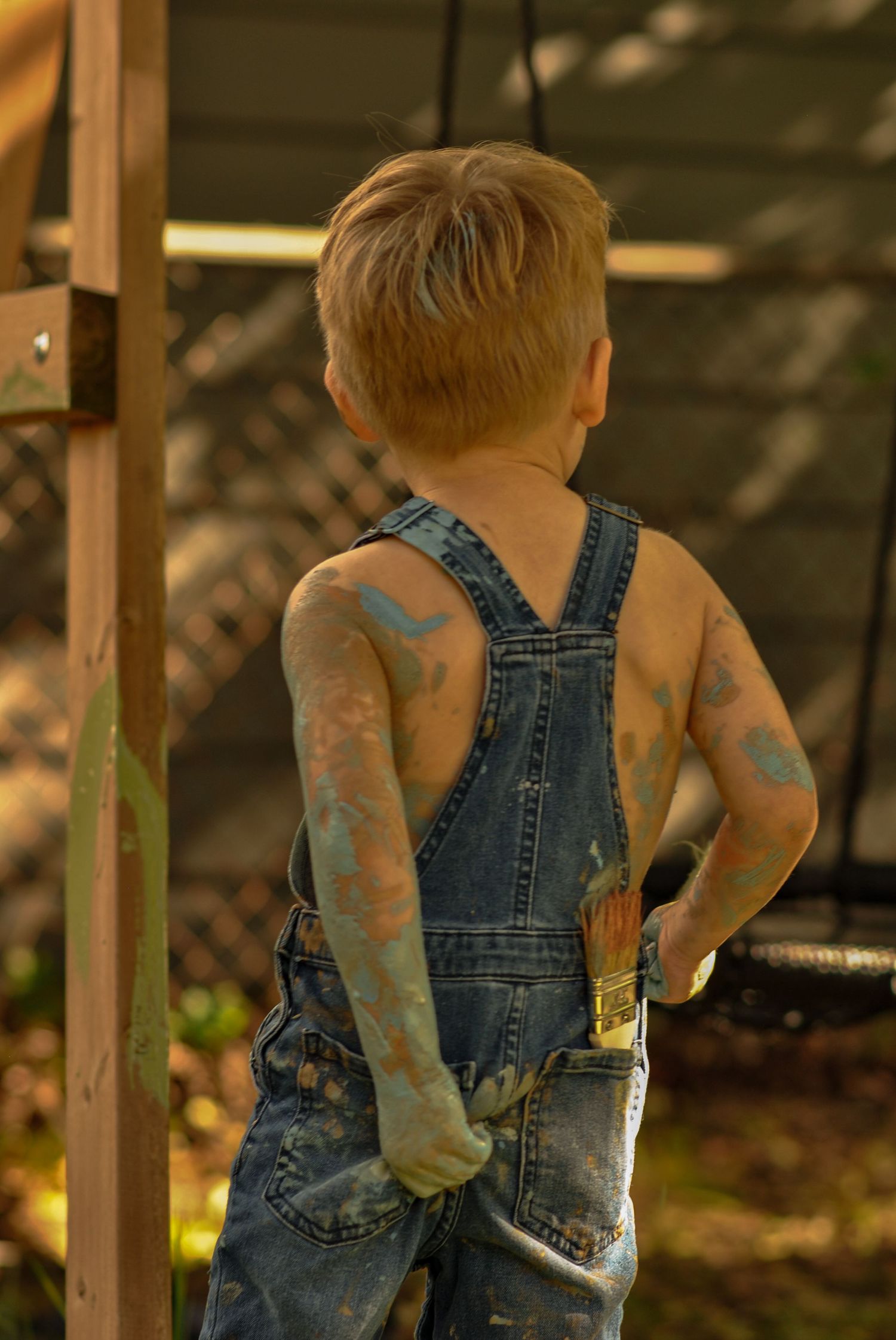 Young boy in denim overalls seen from behind, arms and back covered in blue and green paint, holding a paintbrush outdoors.