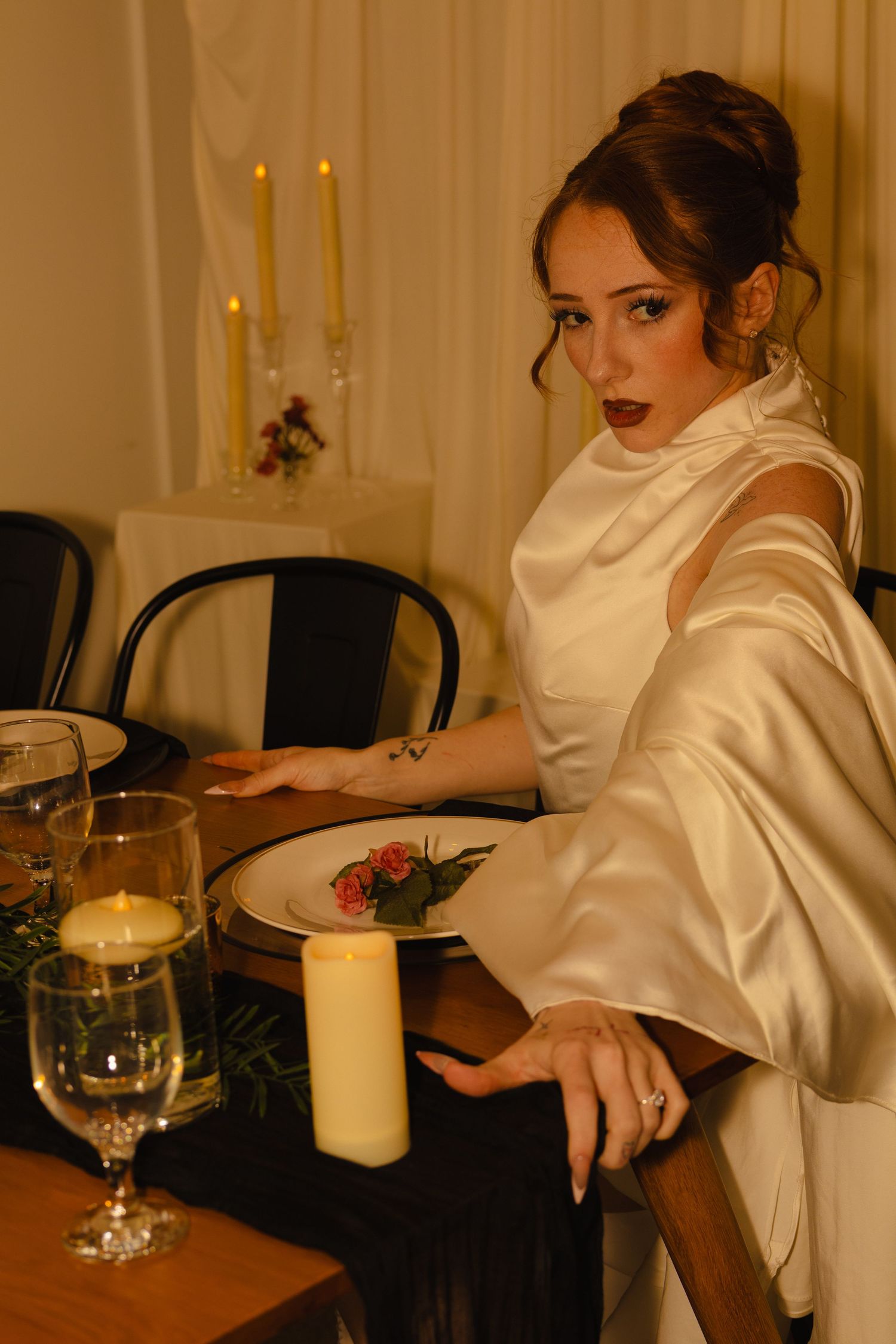 Elegant woman in white satin gown seated at candlelit dinner table with wine glasses and floral plate decor.