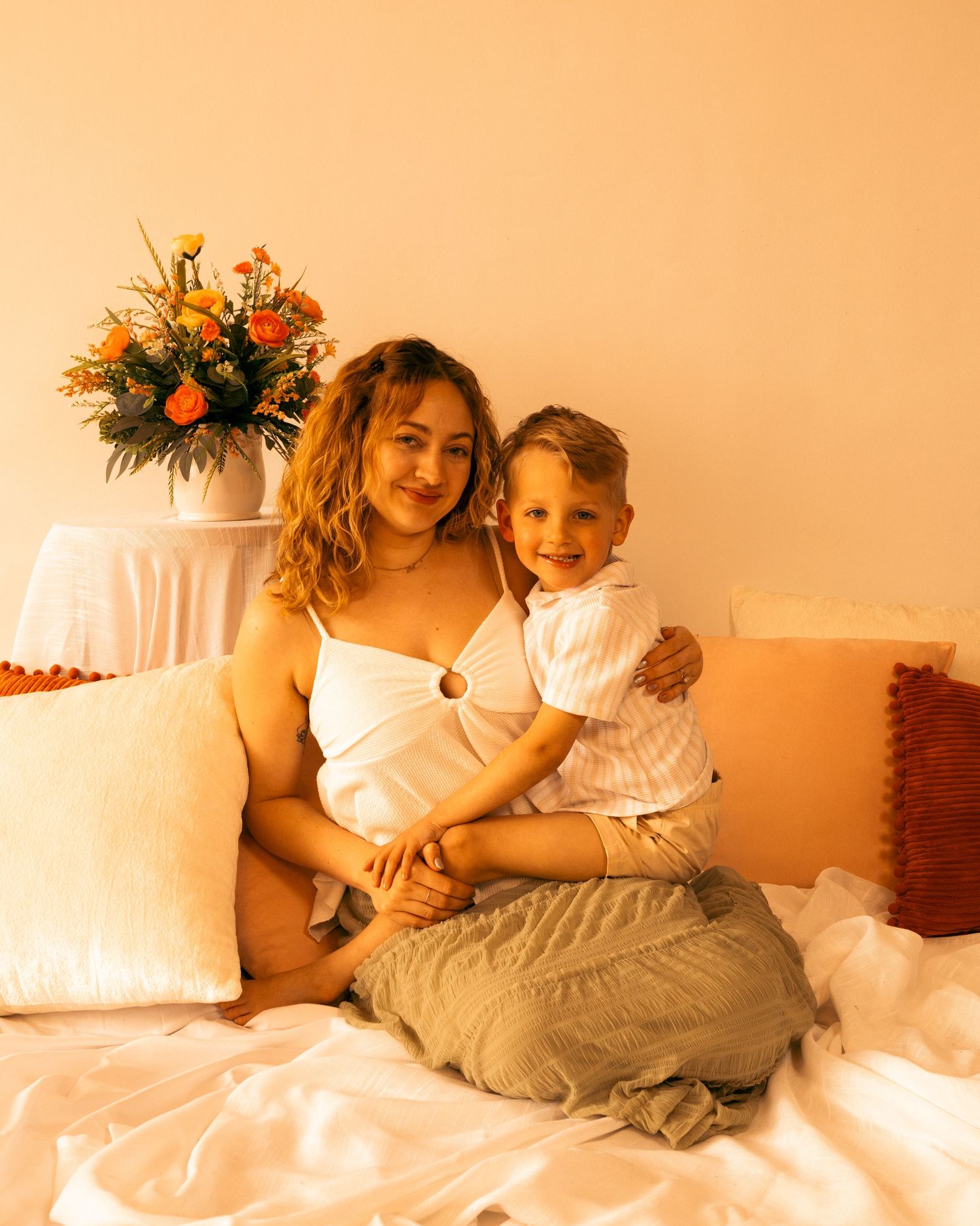 A smiling woman in a white top sits on a bed with a young boy, surrounded by white pillows and orange flowers.