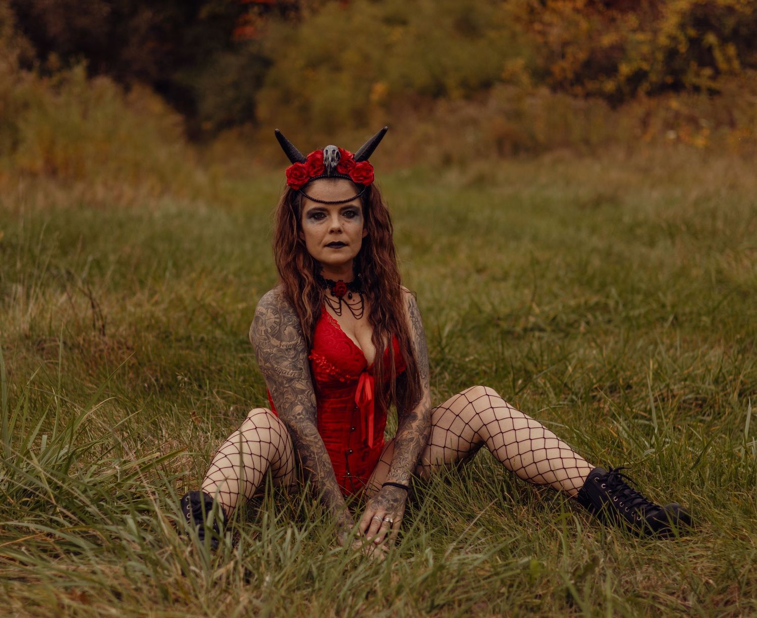 Woman in red devil costume with horns and fishnet stockings sitting in a grassy field for Halloween.