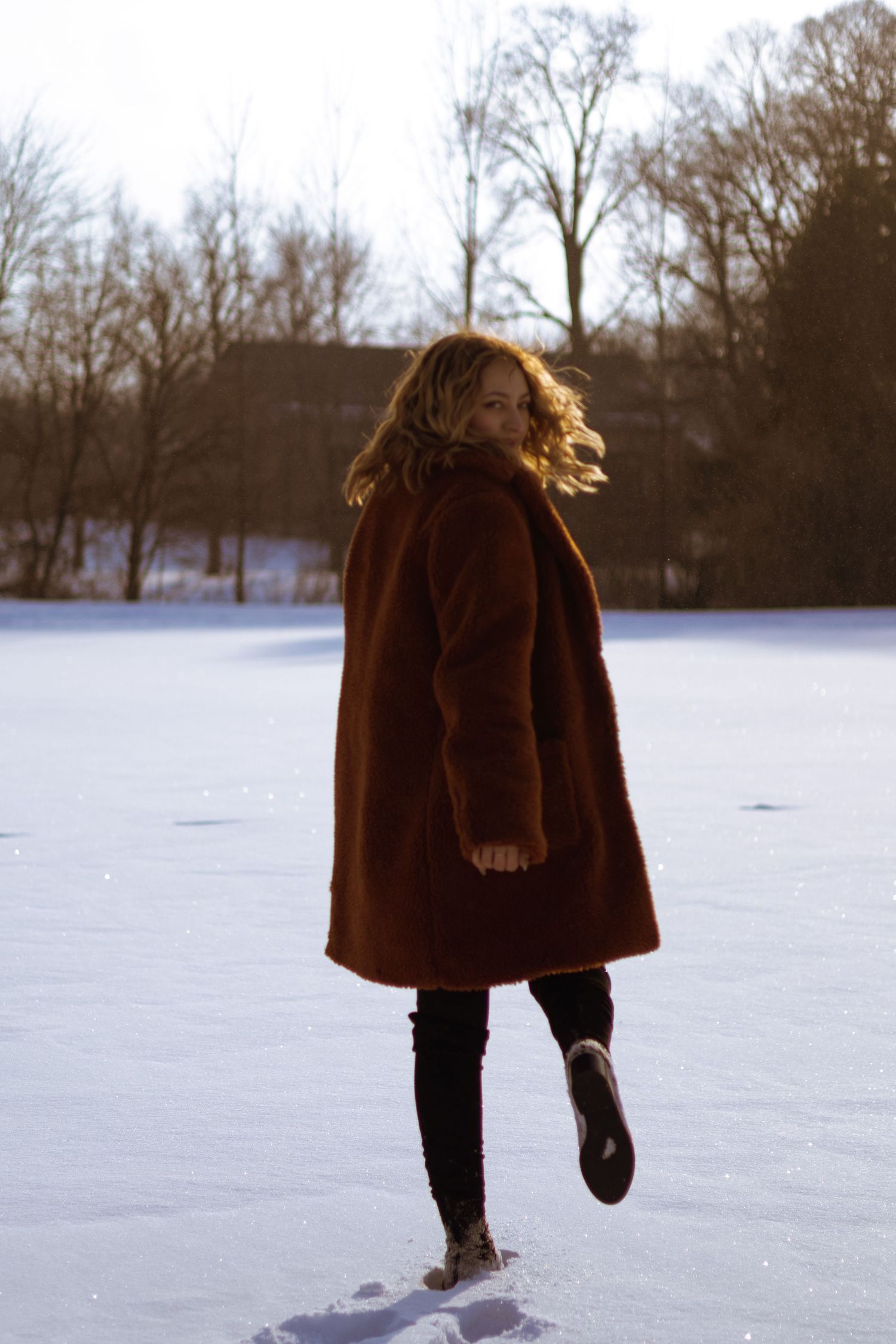 Woman in brown teddy coat walking through snowy park, looking back over shoulder on a bright winter day.