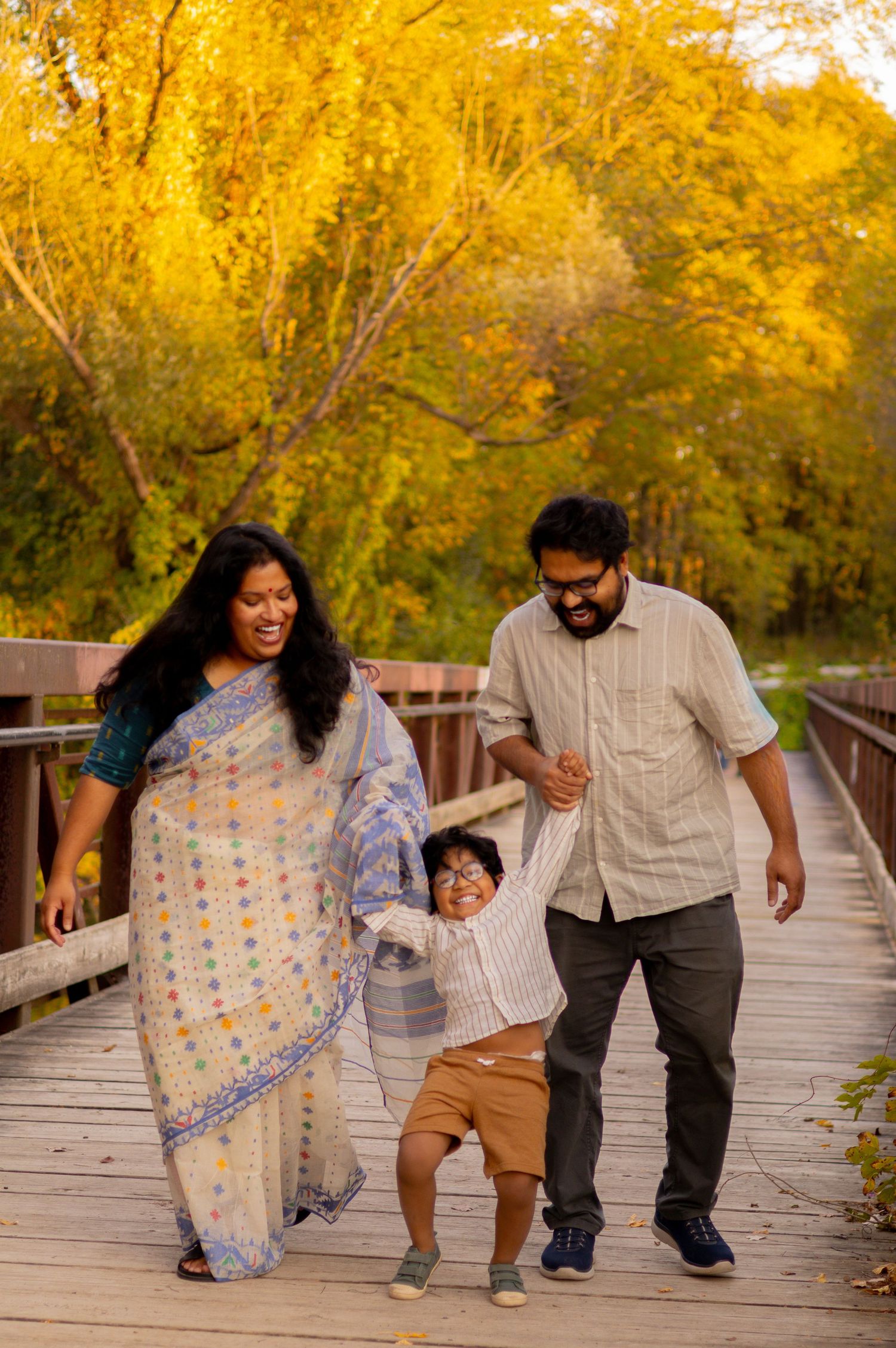 A family walks together holding hands on a wooden boardwalk surrounded by golden fall trees.