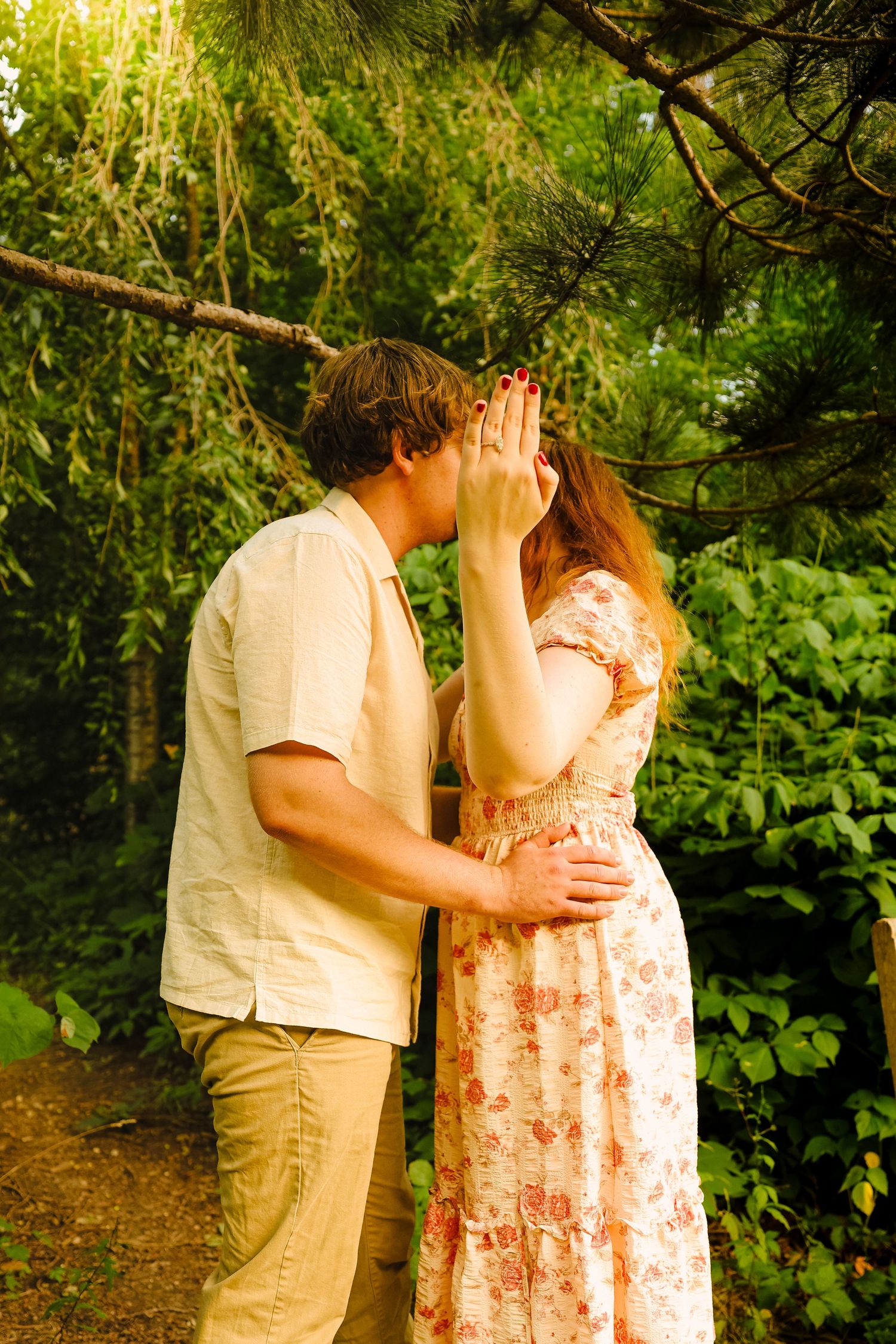 A couple sharing a romantic kiss outdoors in a lush green forest, woman in floral dress covering her face with hands.