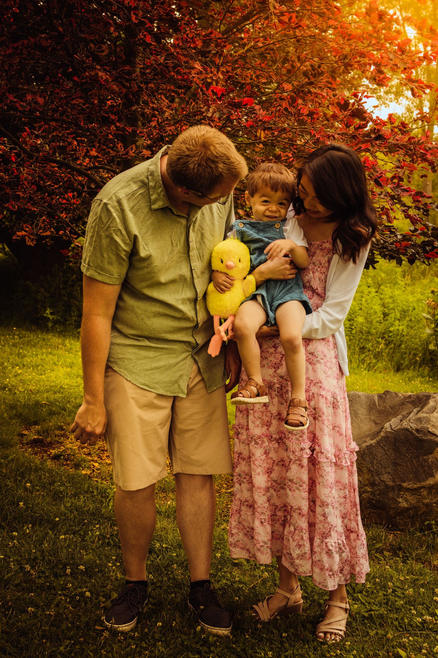 Family of three outdoors in autumn garden, parents lovingly gazing at toddler holding yellow toy duck.