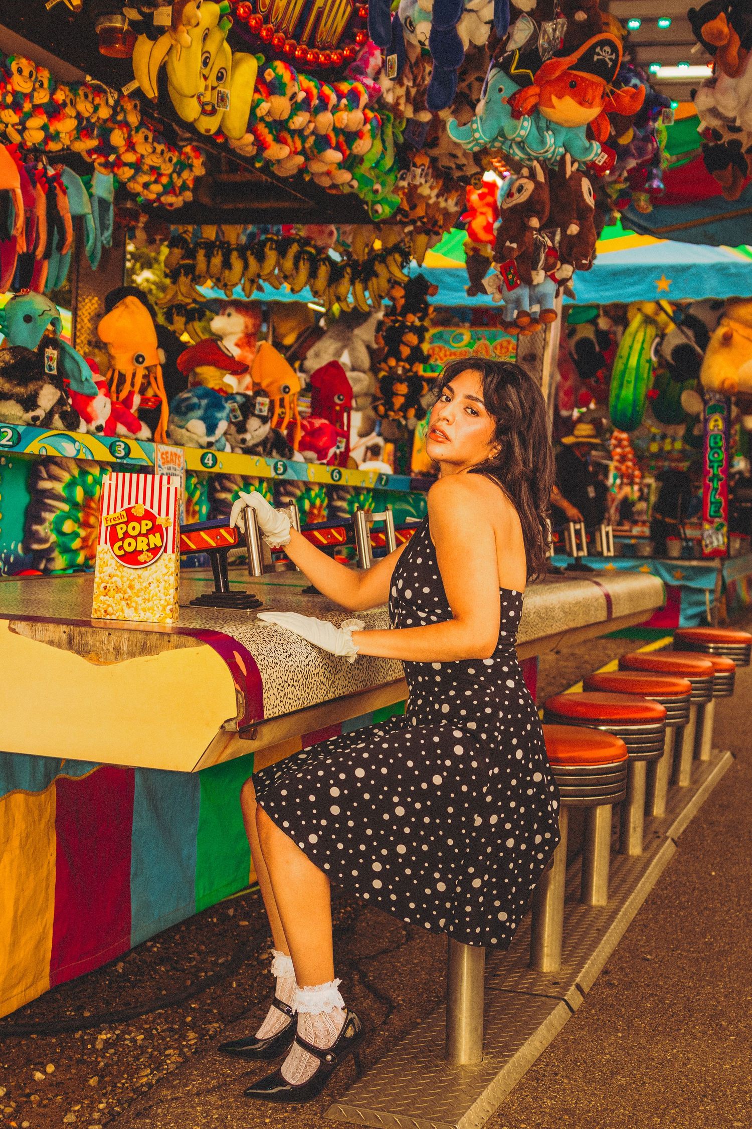 Woman in polka dot dress sitting at colorful carnival game booth with stuffed animal prizes and popcorn.