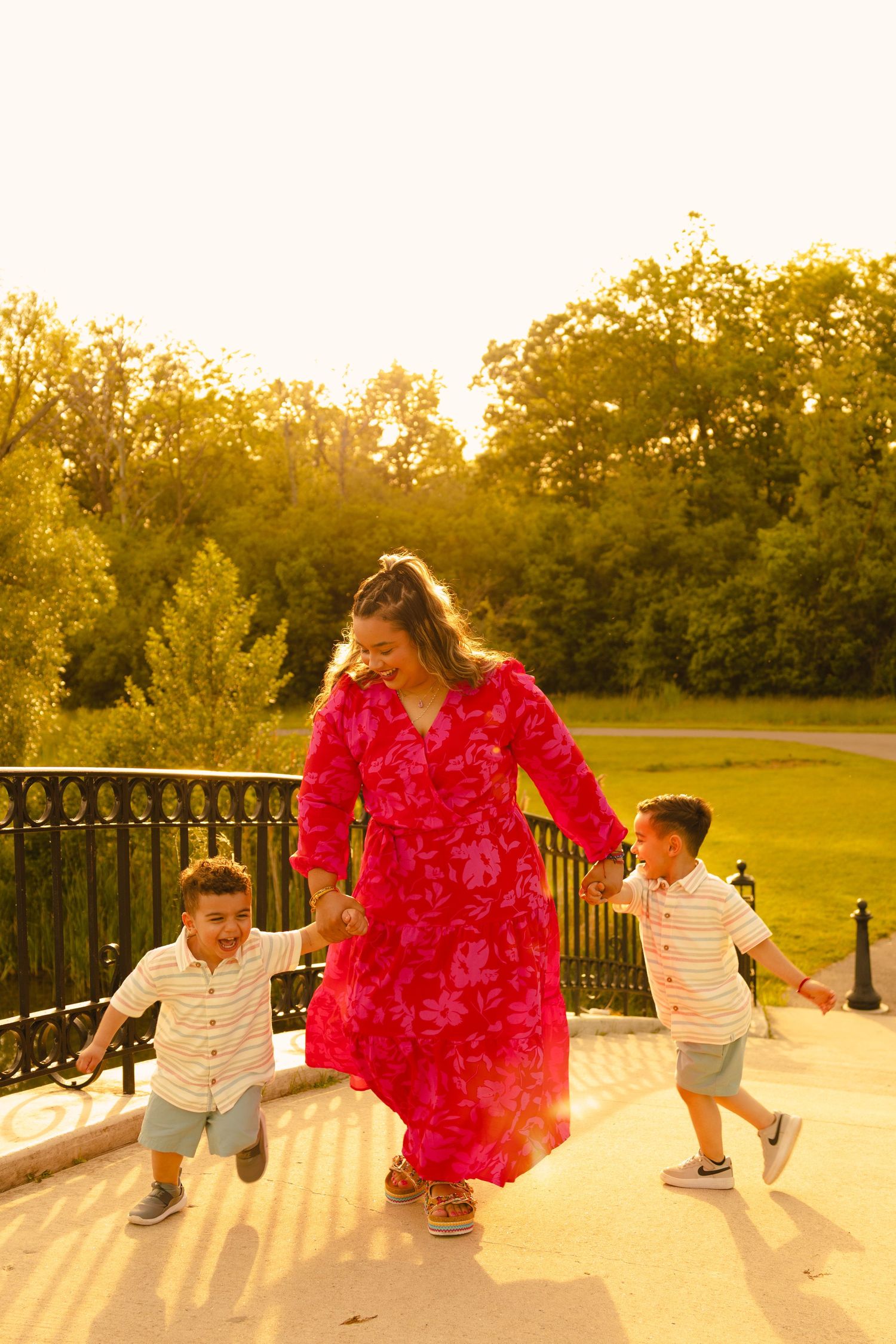 A mother in a red floral dress holds hands with her two young sons on a bridge in a sunlit park.