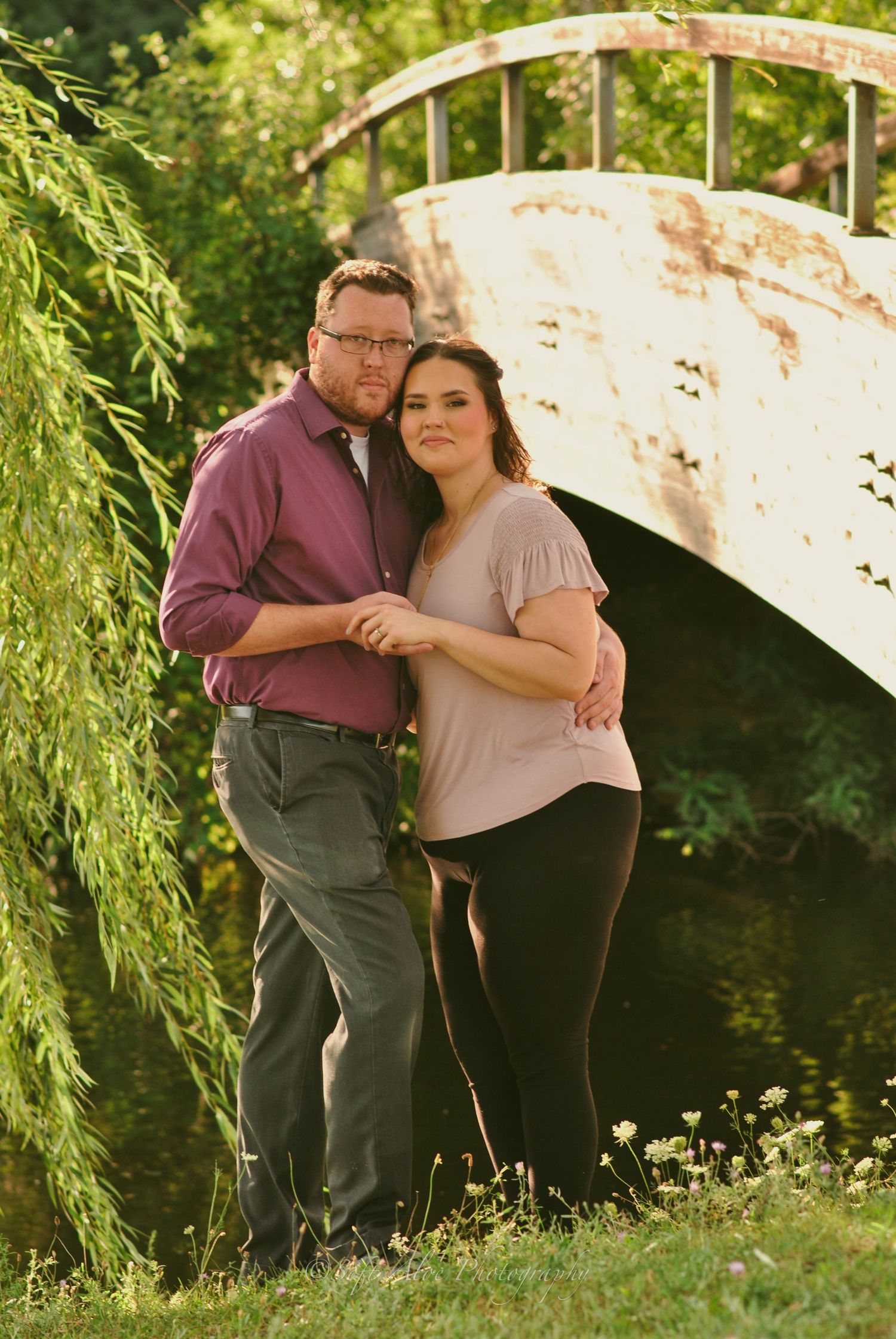 A loving couple embraces under willow trees near a rustic bridge in a garden setting.