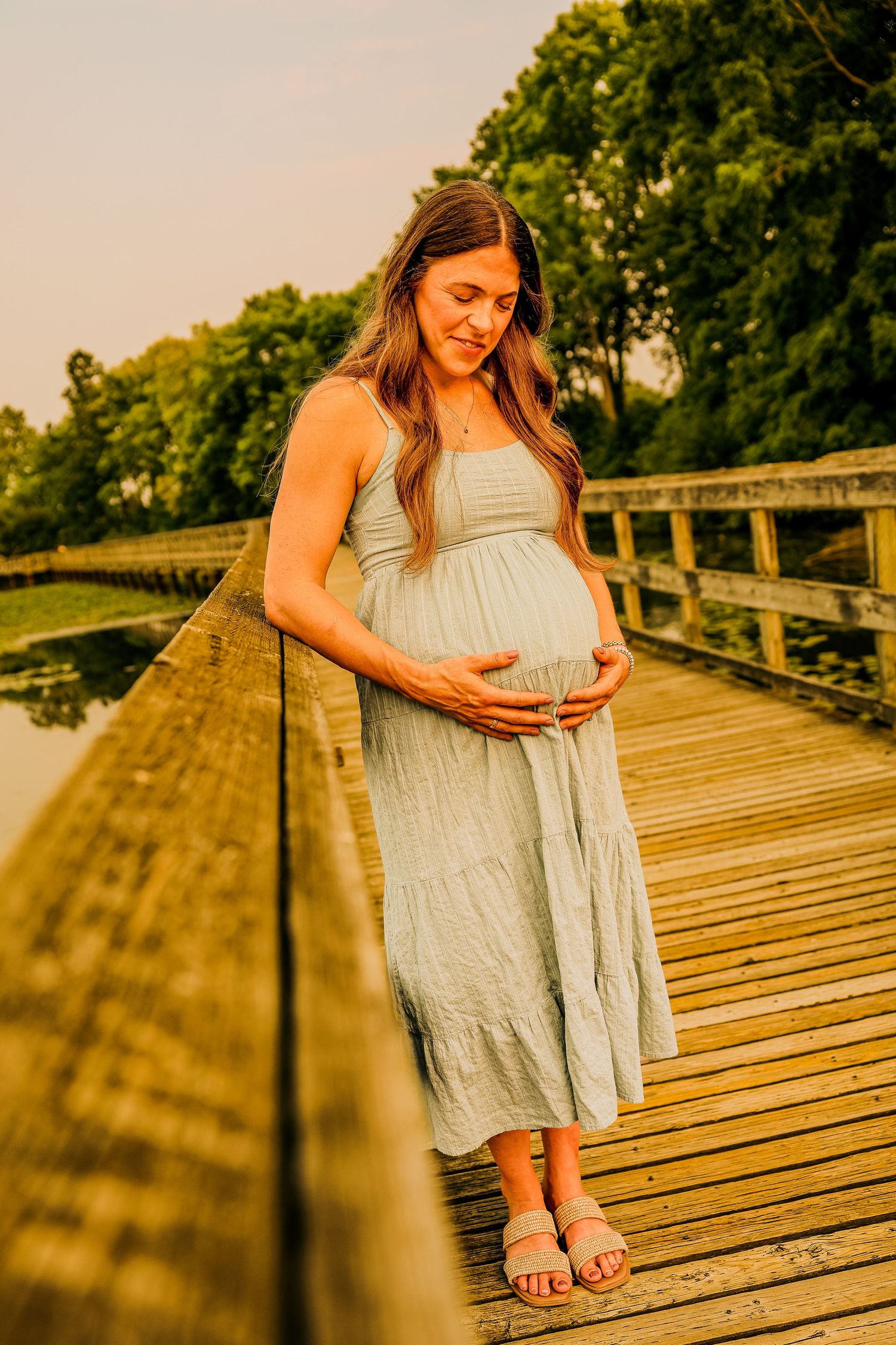 Pregnant woman in sage green dress cradling her baby bump on a wooden bridge at golden hour.