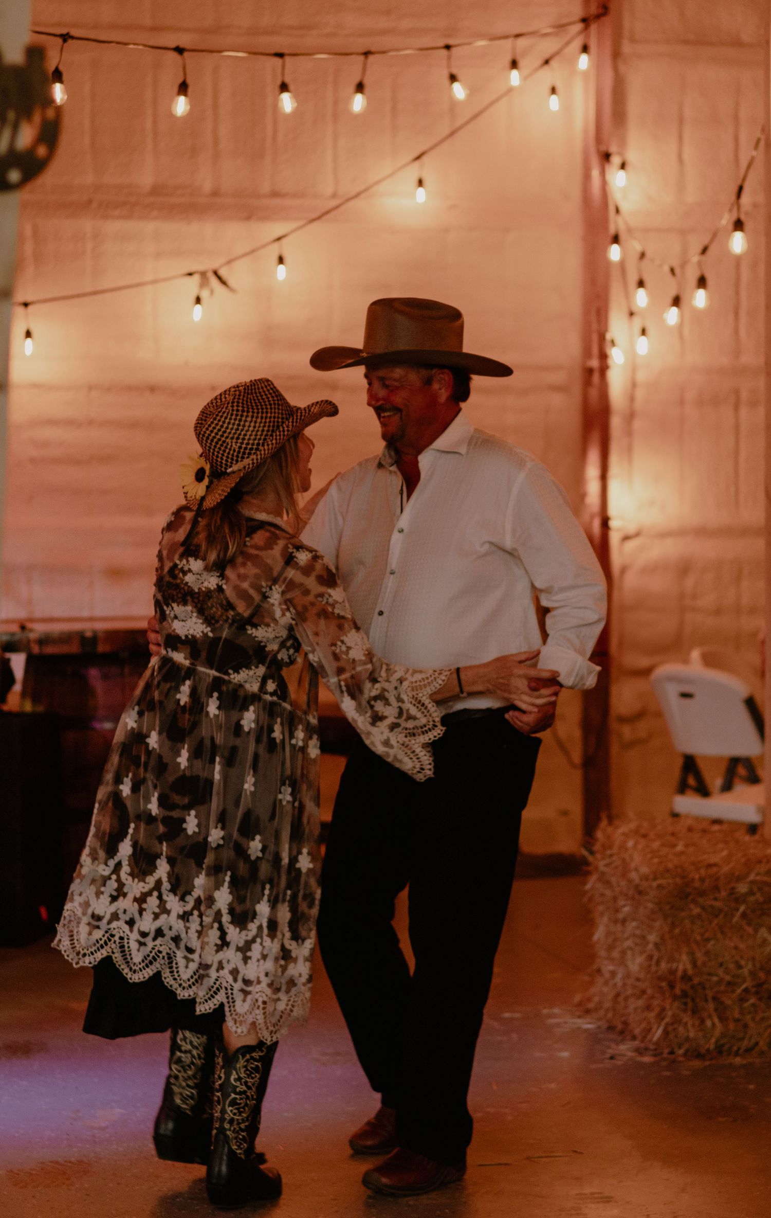 A couple in western attire dancing together in a rustic barn decorated with string lights and hay bales.