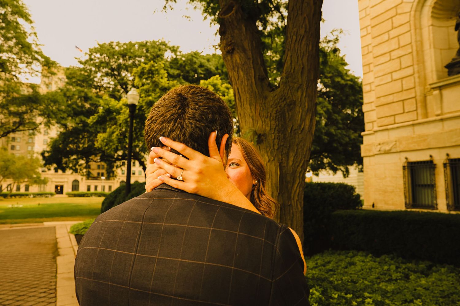Couple embracing near a tree in golden hour light, woman's hand with ring visible on man's back.