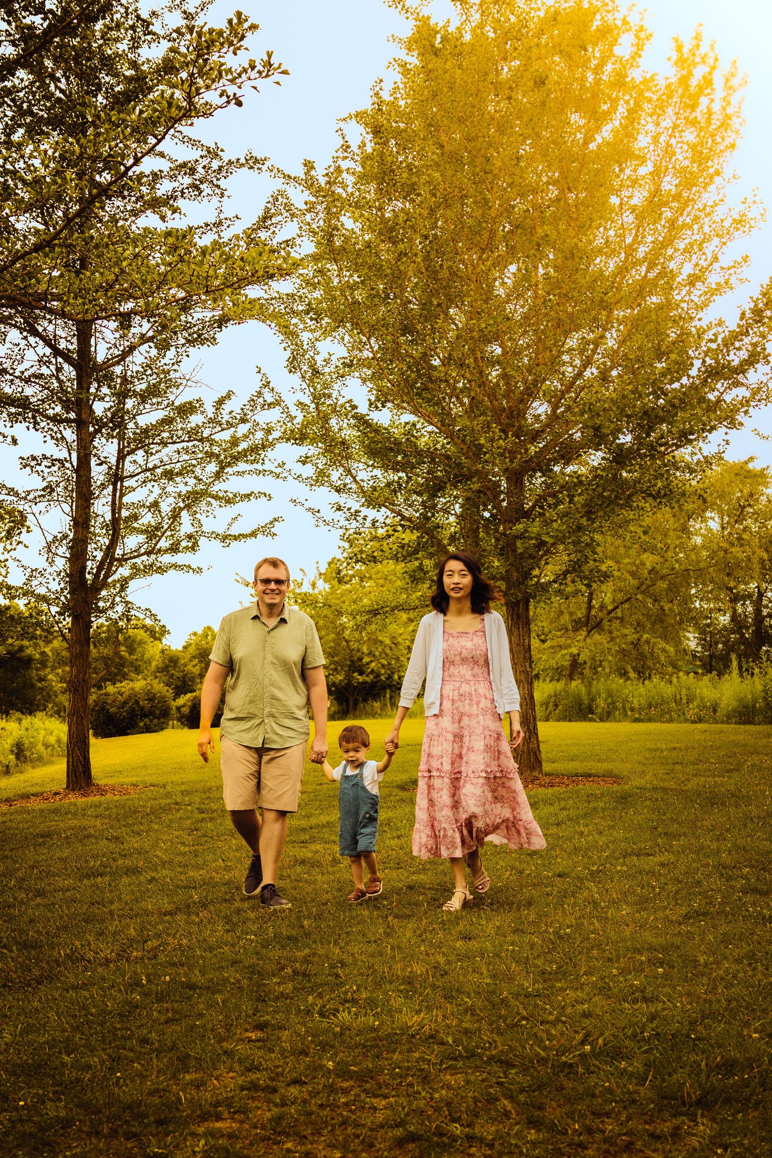 Family of three walking hand-in-hand through a sunny green park during golden hour.