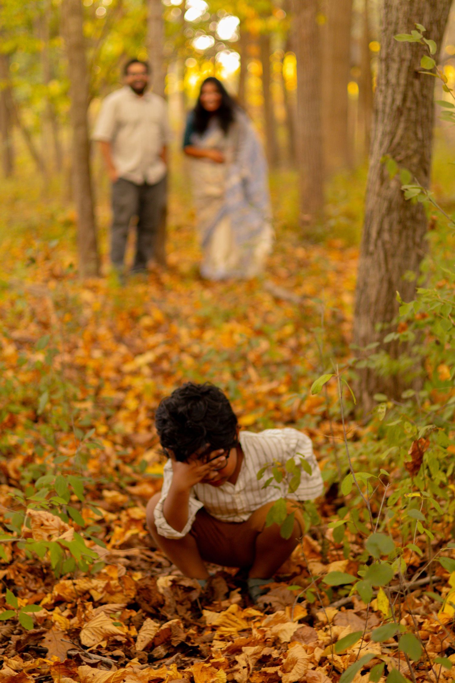 A child plays in fallen autumn leaves while parents watch from a distance.