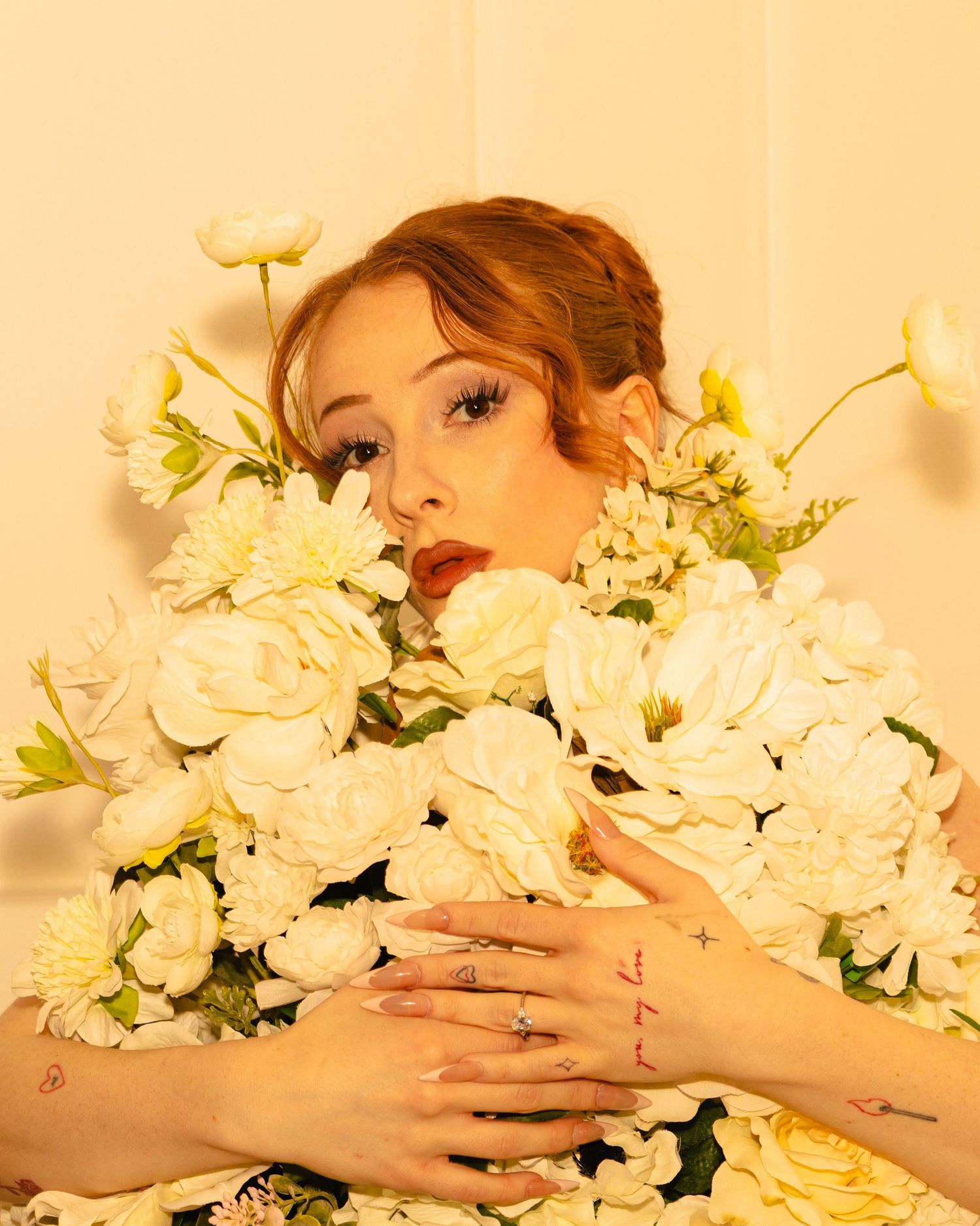 A woman with red hair embraces white flowers including ranunculus and lilies in a soft, dreamy floral portrait.