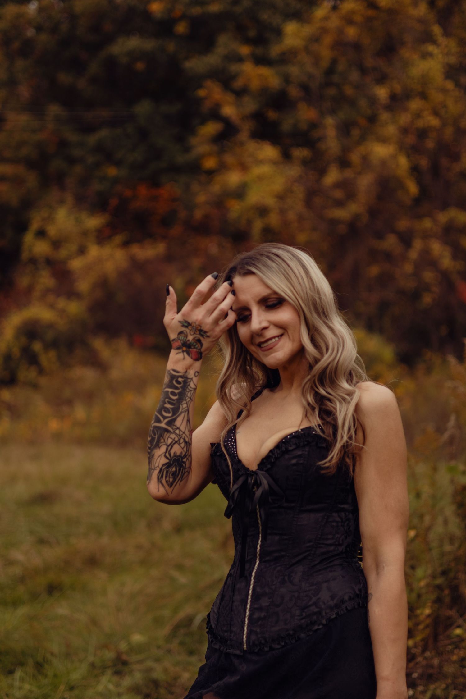 Blonde woman in black lace dress with tattoos poses outdoors among autumn foliage, smiling softly.
