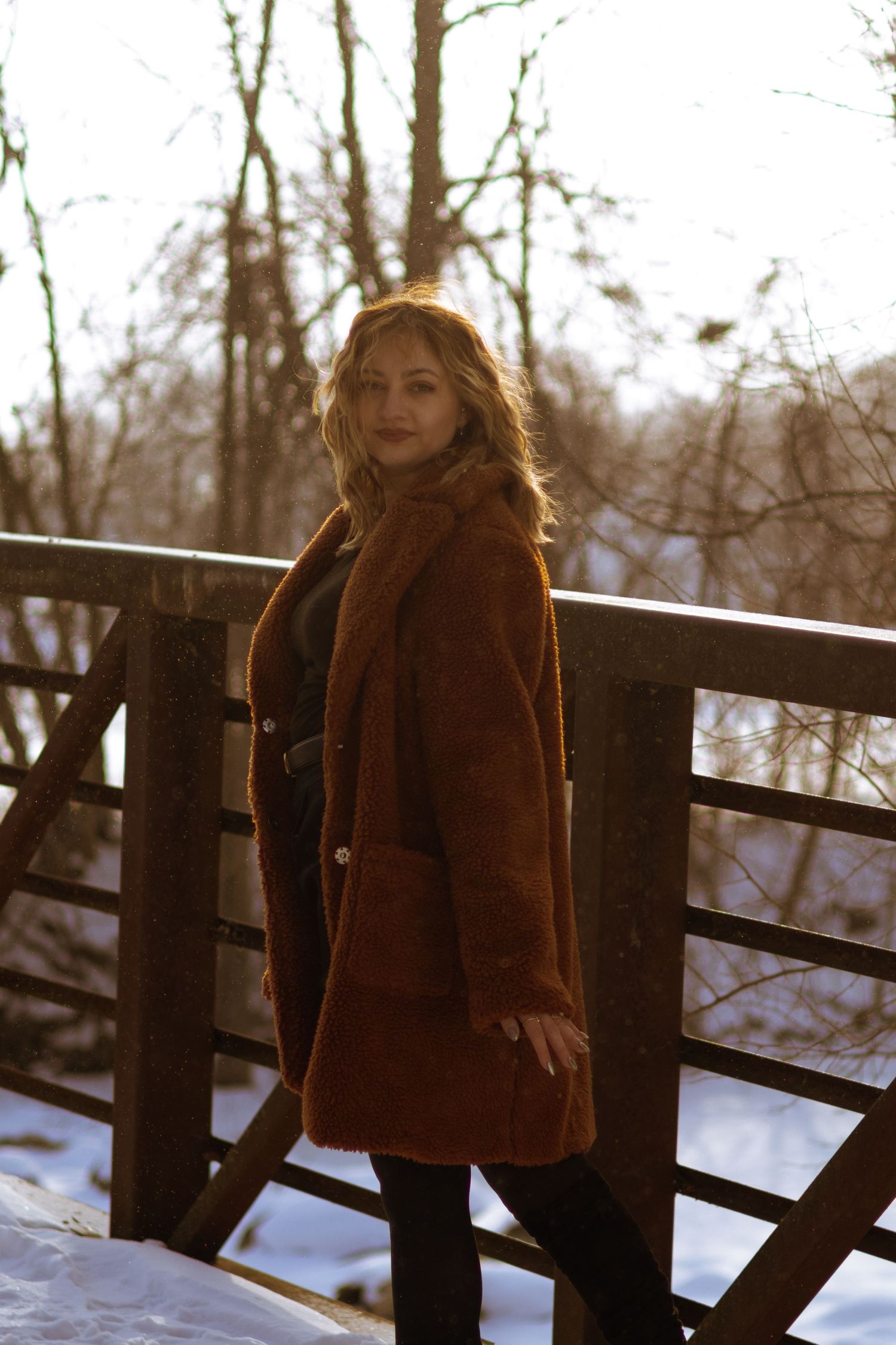 Woman in brown teddy coat standing on snowy wooden bridge at sunset with bare trees in background.