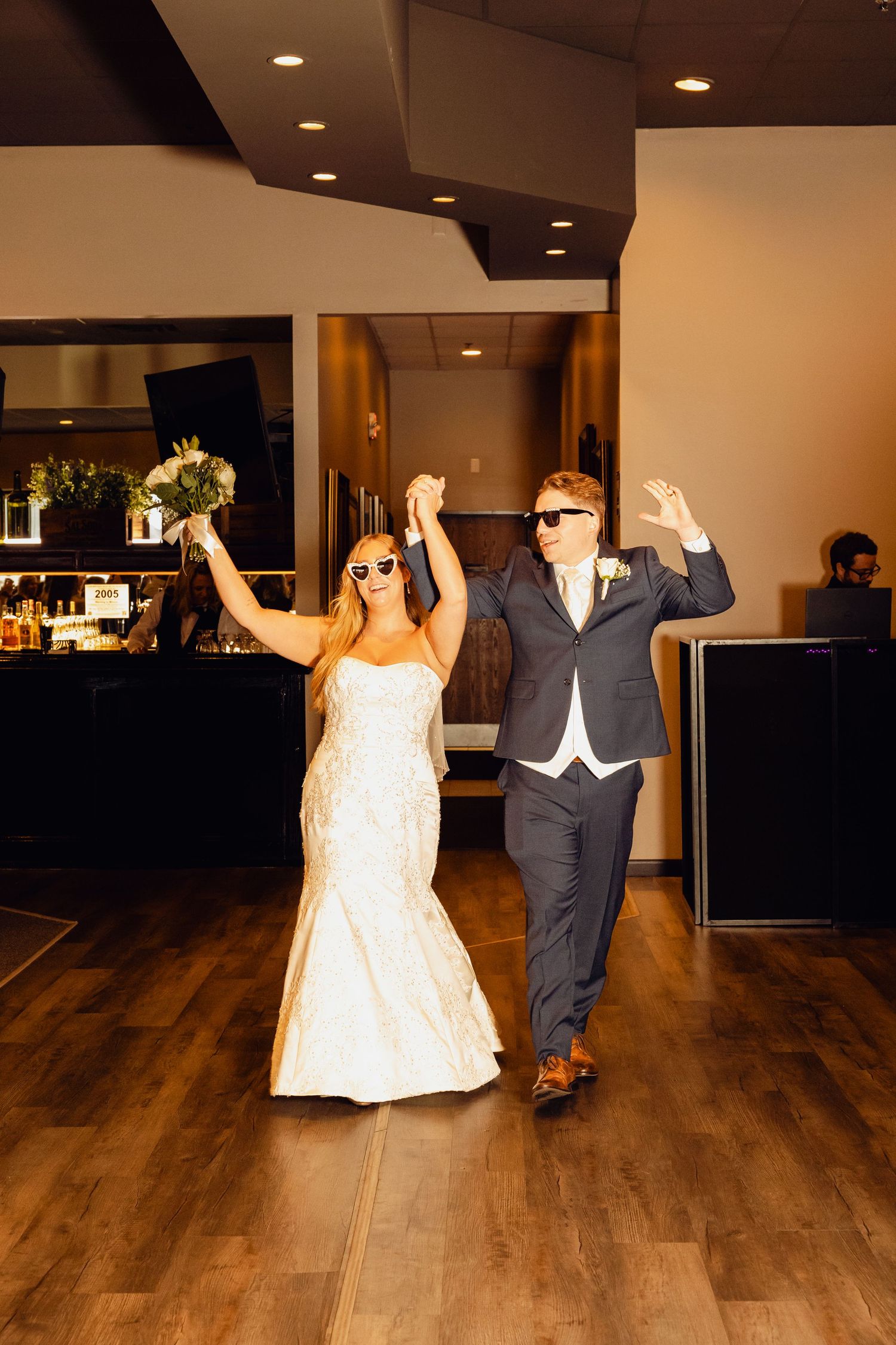Smiling bride in a white gown and groom in a navy suit, both wearing sunglasses, raise their arms as they enter their wedding reception.
