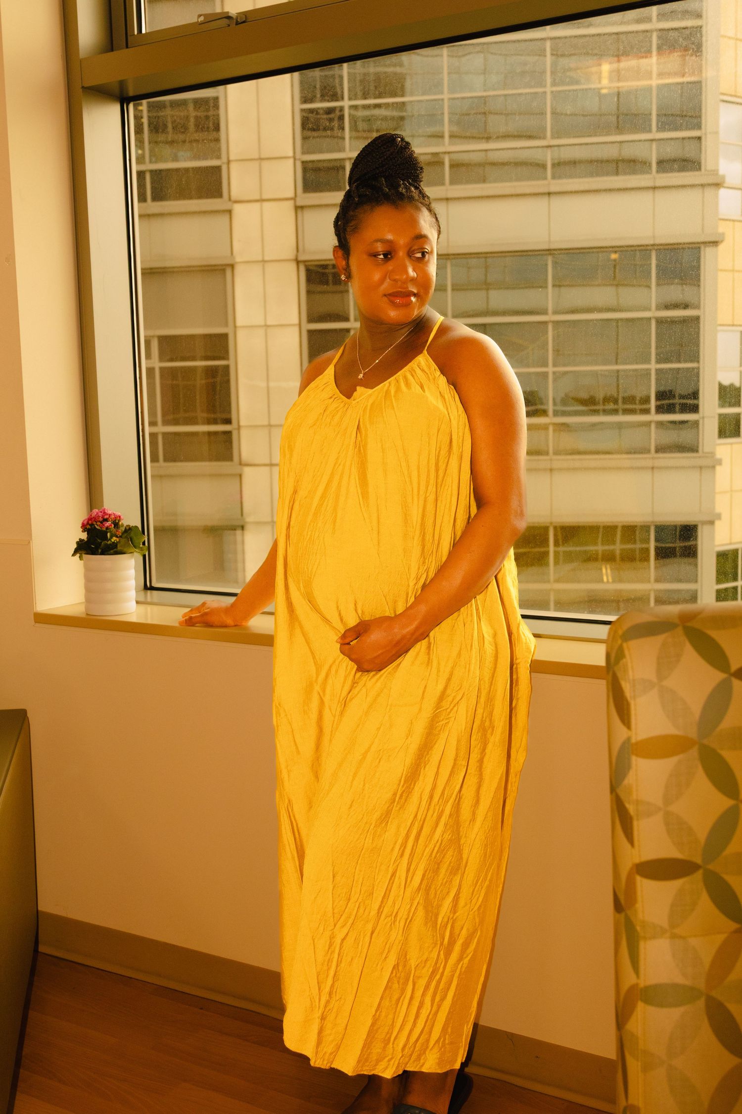 Pregnant woman in yellow maxi dress standing by window in modern building interior.
