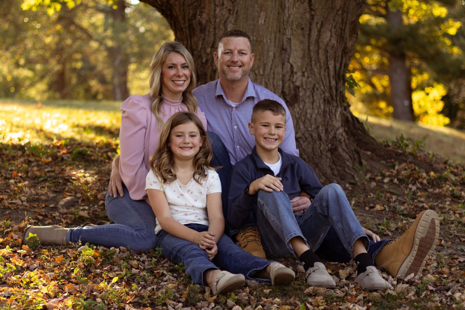 Family relaxes together at base of large tree trunk in autumn forest setting.