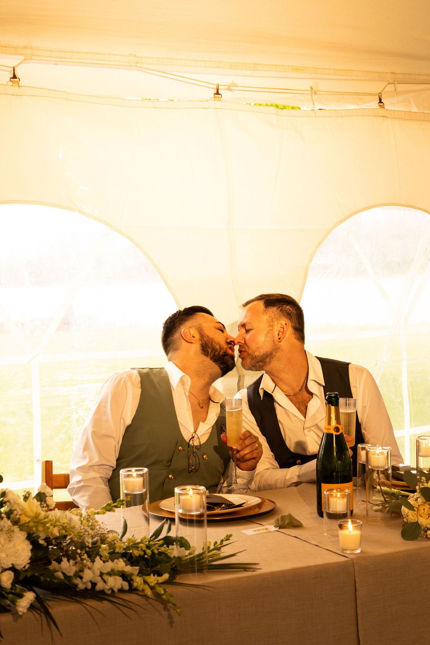 Two grooms share a kiss at their wedding reception table with champagne glasses and floral centerpieces.