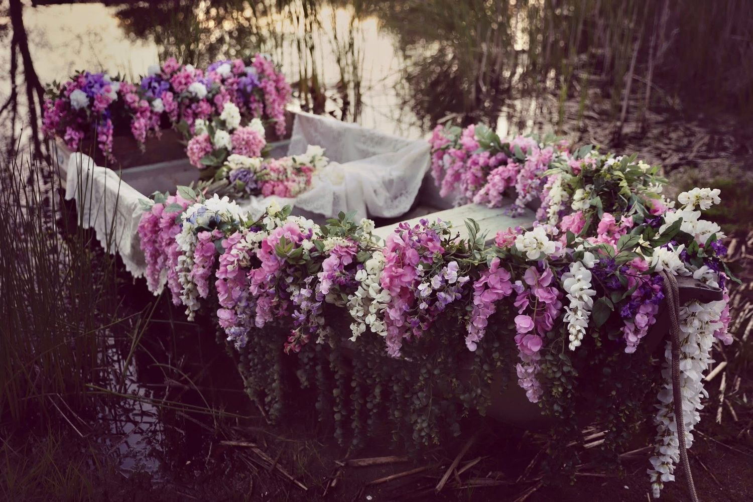 A wooden boat overflowing with lush pink and purple floral arrangements floating on a calm pond.