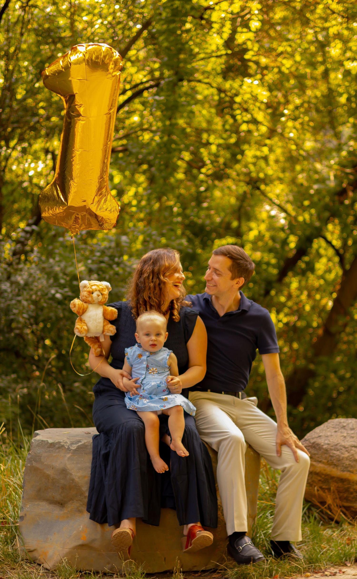 Family celebrating baby's first birthday outdoors, with gold number 1 balloon and teddy bear, seated on rocks in a lush park.