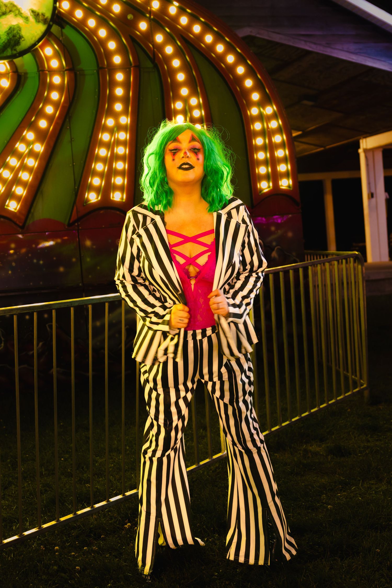 Person in black and white striped suit with green wig poses at night carnival with illuminated carousel backdrop.
