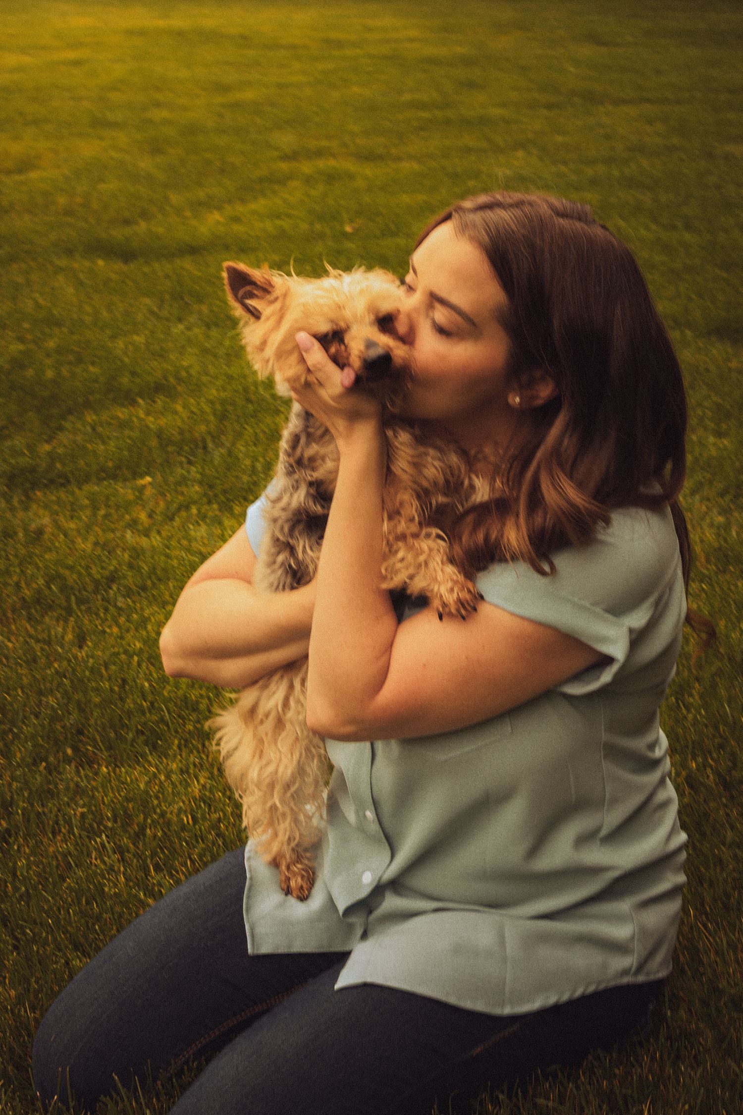 Woman in mint top kisses small Yorkshire Terrier puppy while sitting on lush green grass outdoors.
