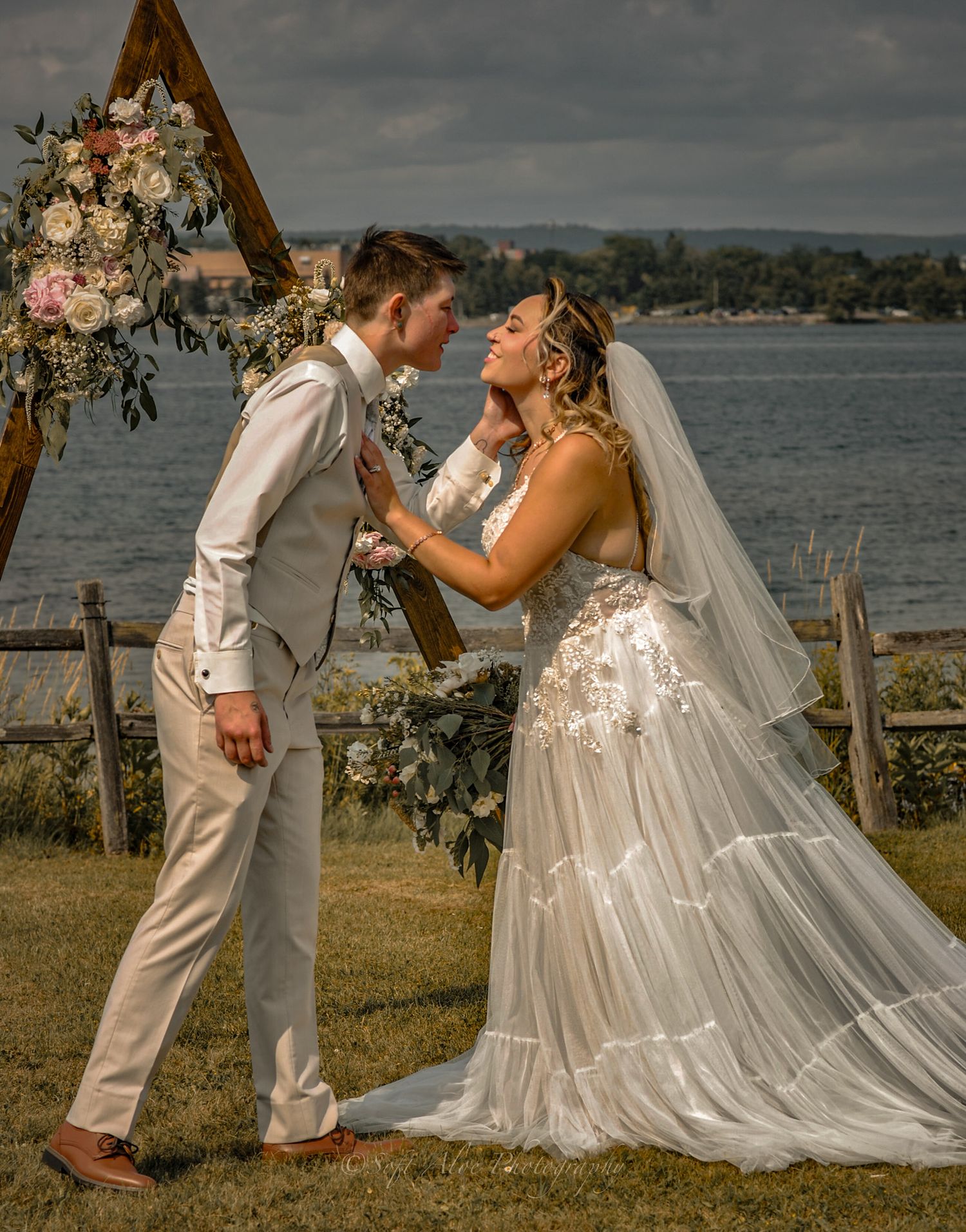 Wedding couple sharing a romantic moment under a wooden arch by the waterfront.