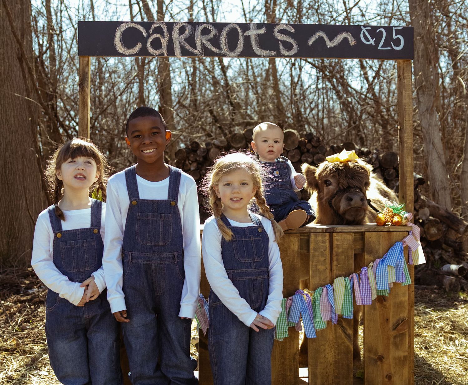 Four children in denim overalls pose at a rustic carrot stand with a chalkboard sign reading Carrots 25 cents.