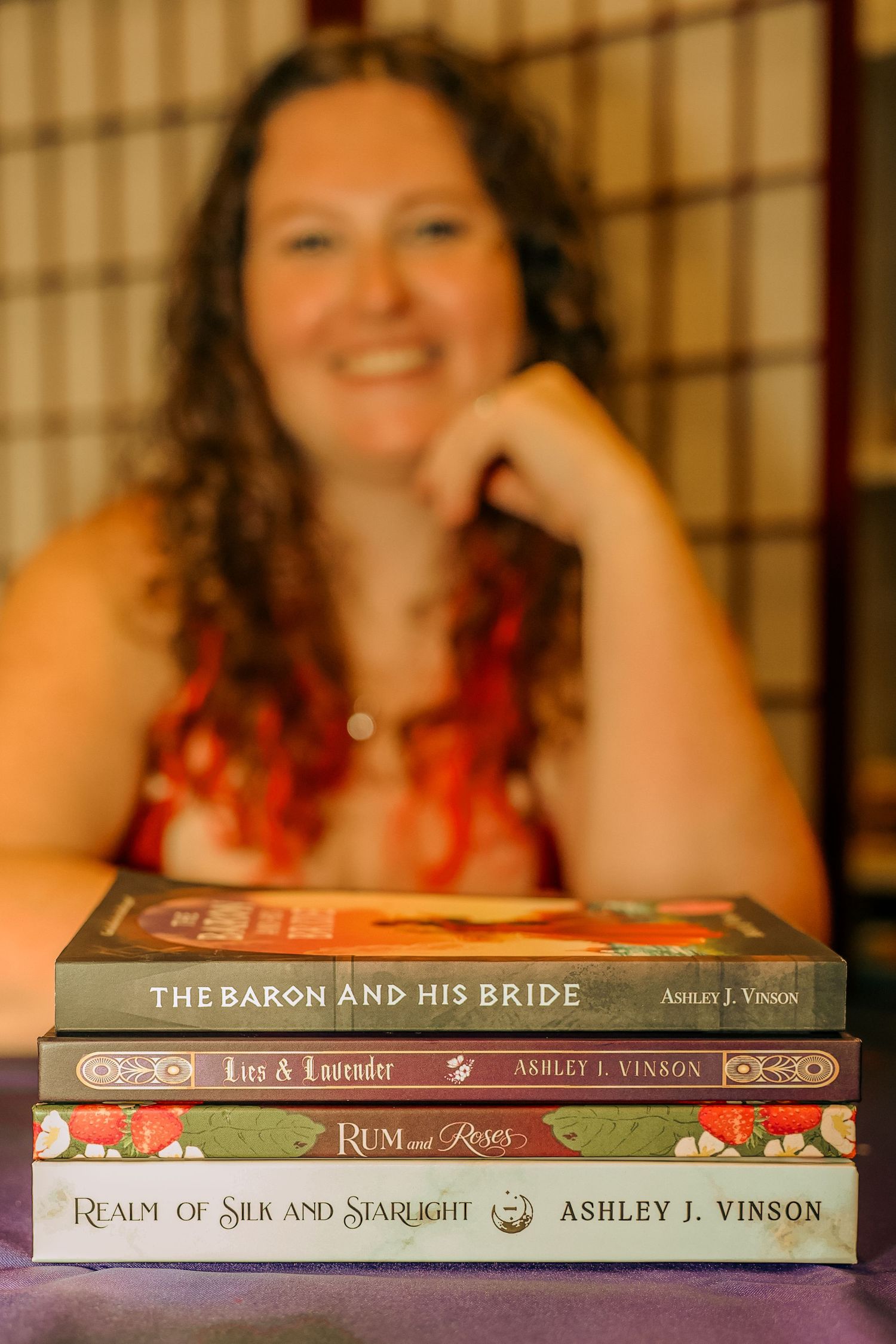 Author Ashley J. Vinson smiling behind a stack of her four published books on a purple table.