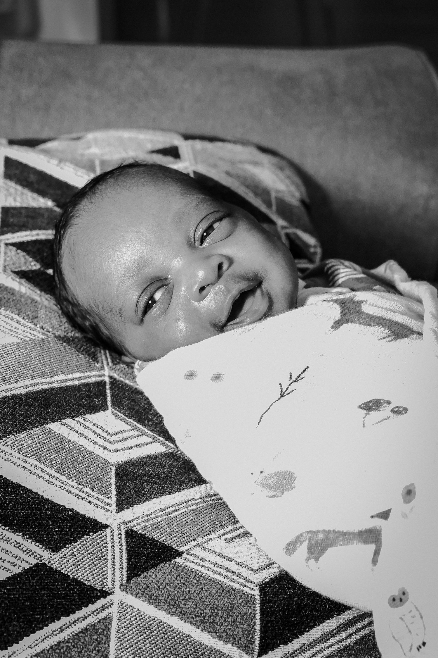 Black and white photo of a smiling newborn baby lying on a patterned blanket with an animal print pillow.