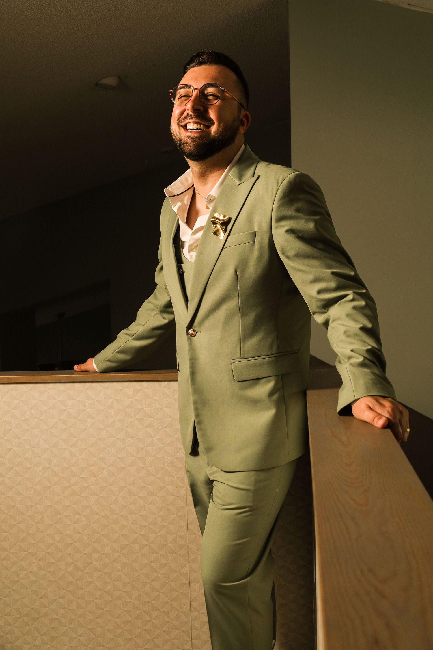 Smiling man in sage green suit with boutonniere, wearing glasses, leaning on railing in warm indoor lighting.