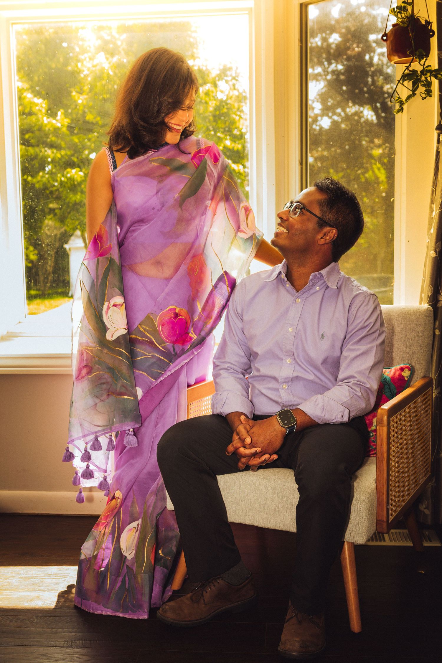 A couple shares a tender moment, woman in purple floral saree stands beside seated man in lavender shirt by sunlit window.