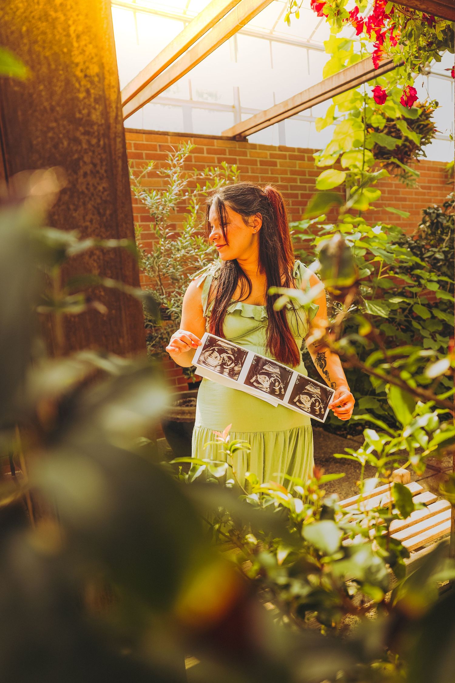 Pregnant woman in green dress holding ultrasound photos in a sunlit garden greenhouse with lush plants and red flowers.