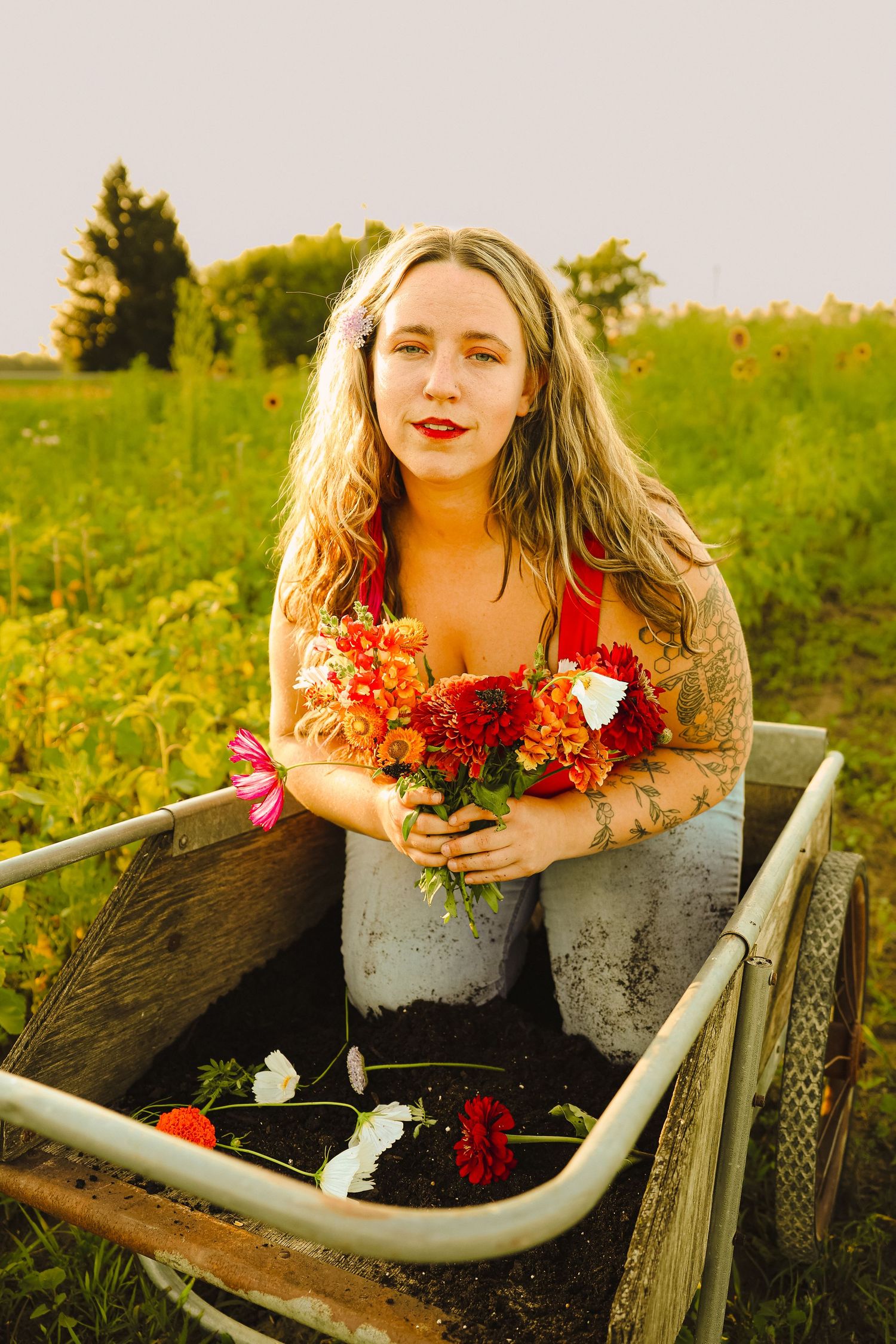 Blonde woman with tattoos holding colorful flowers while sitting in a rustic wooden wheelbarrow in a lush green field.